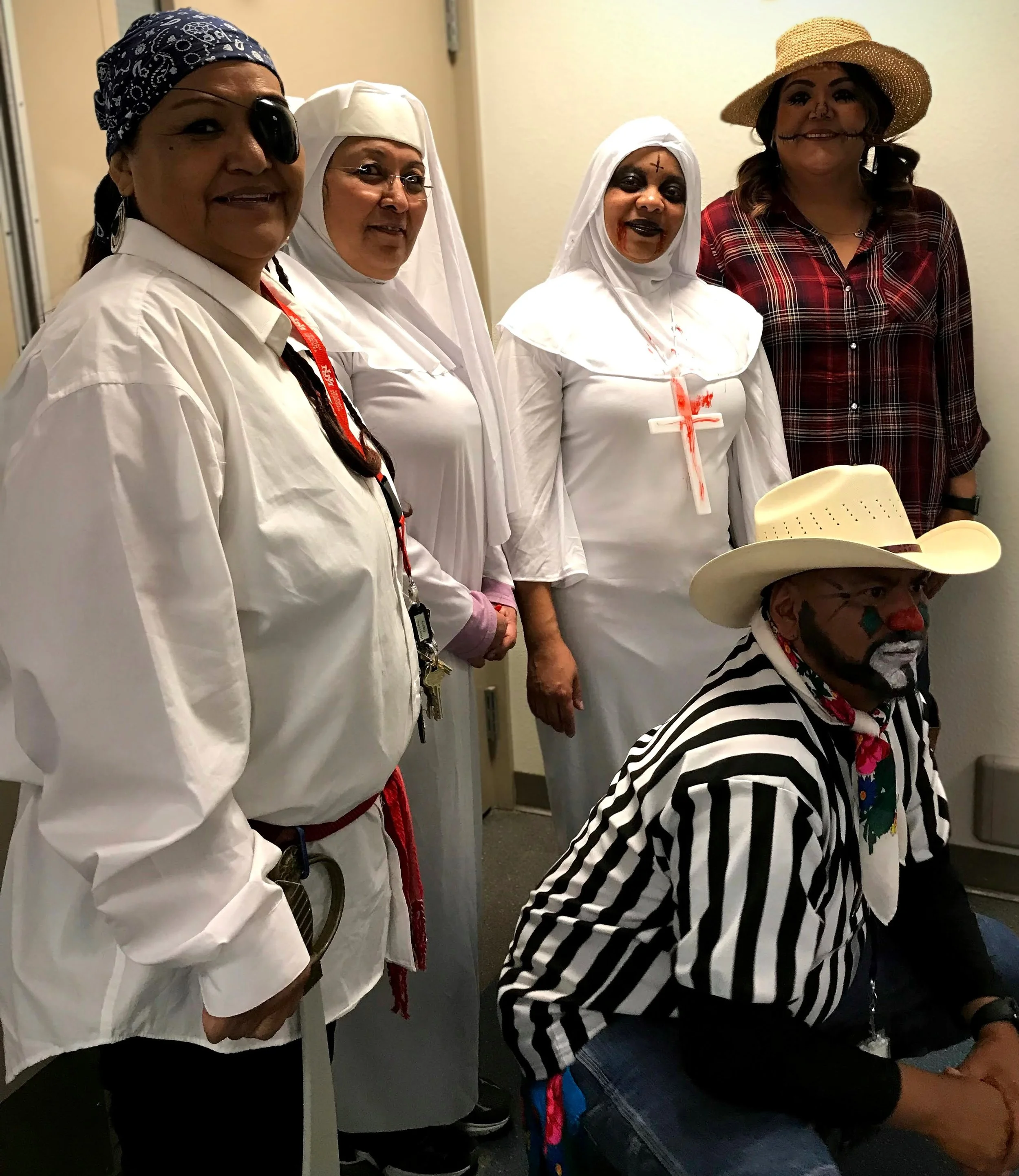 Four women dressed in costumes pose together at the back of a room. The woman on the left wears a pirate costume with an eye patch. The second woman wears a nun costume with glasses. The third woman is dressed as a zombie nun with blood on her face. 