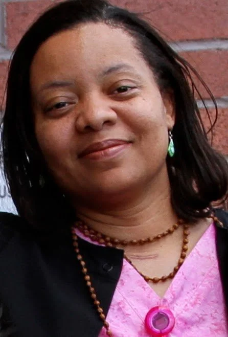 A woman with dark hair and earrings, smiling in front of a brick wall, wearing a pink and black top with jewelry.