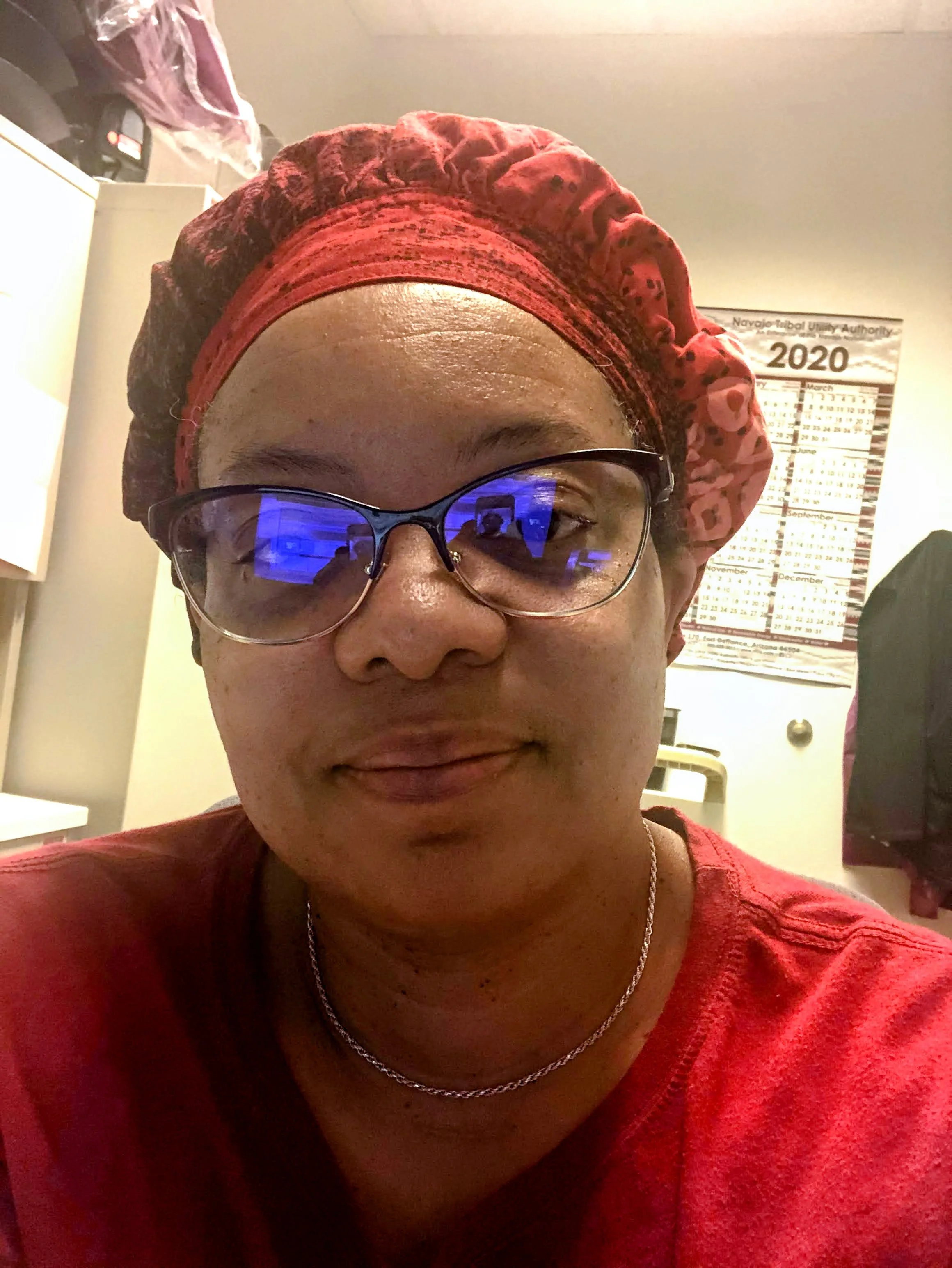 A woman wearing glasses, a red headscarf, and a red shirt. She is smiling slightly and is indoors, with a calendar on the wall and various items in the background.