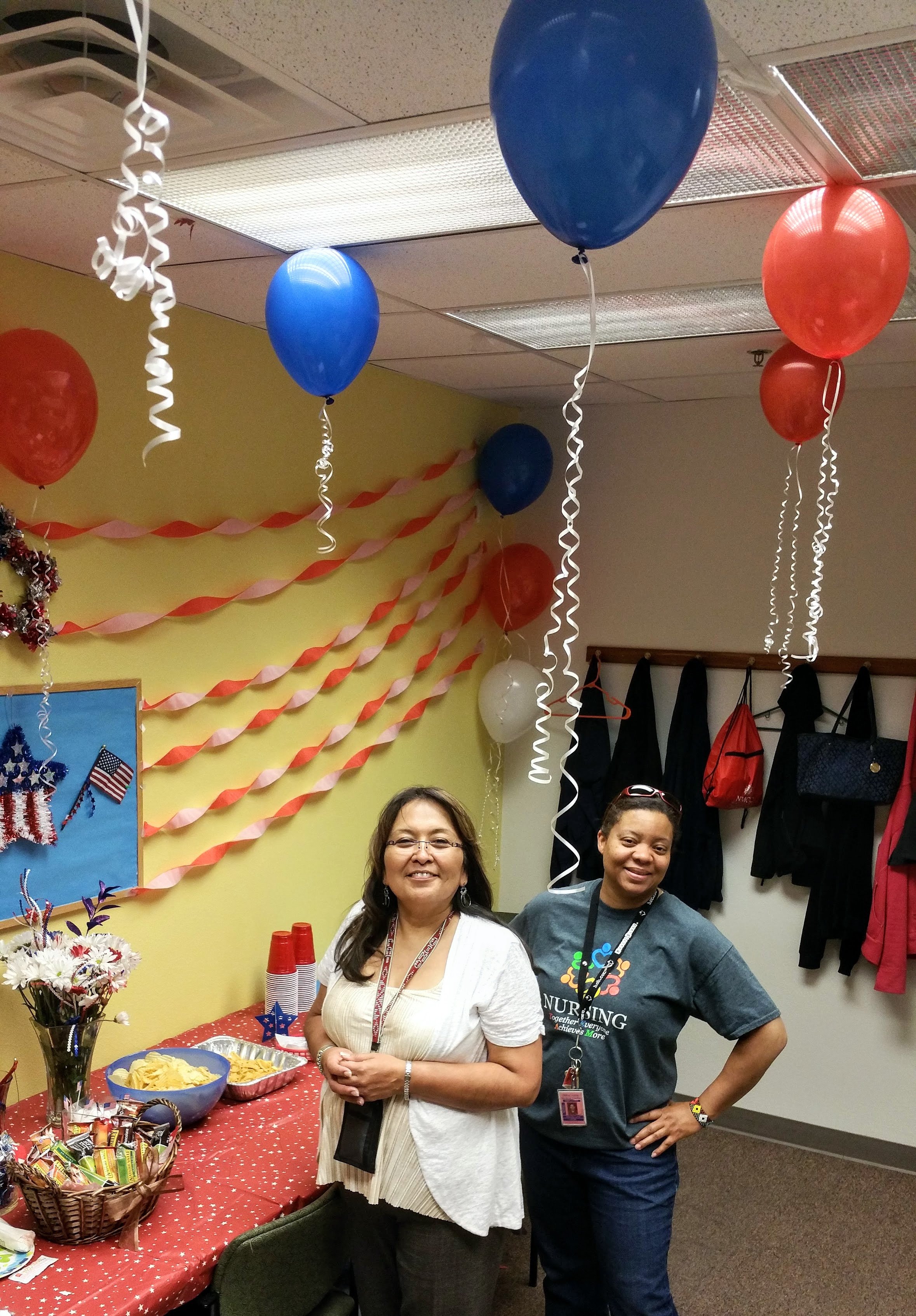 Two women smiling in a decorated room with red, white, and blue balloons and streamers, standing behind a table with snacks, flowers, and cups for a festive celebration.