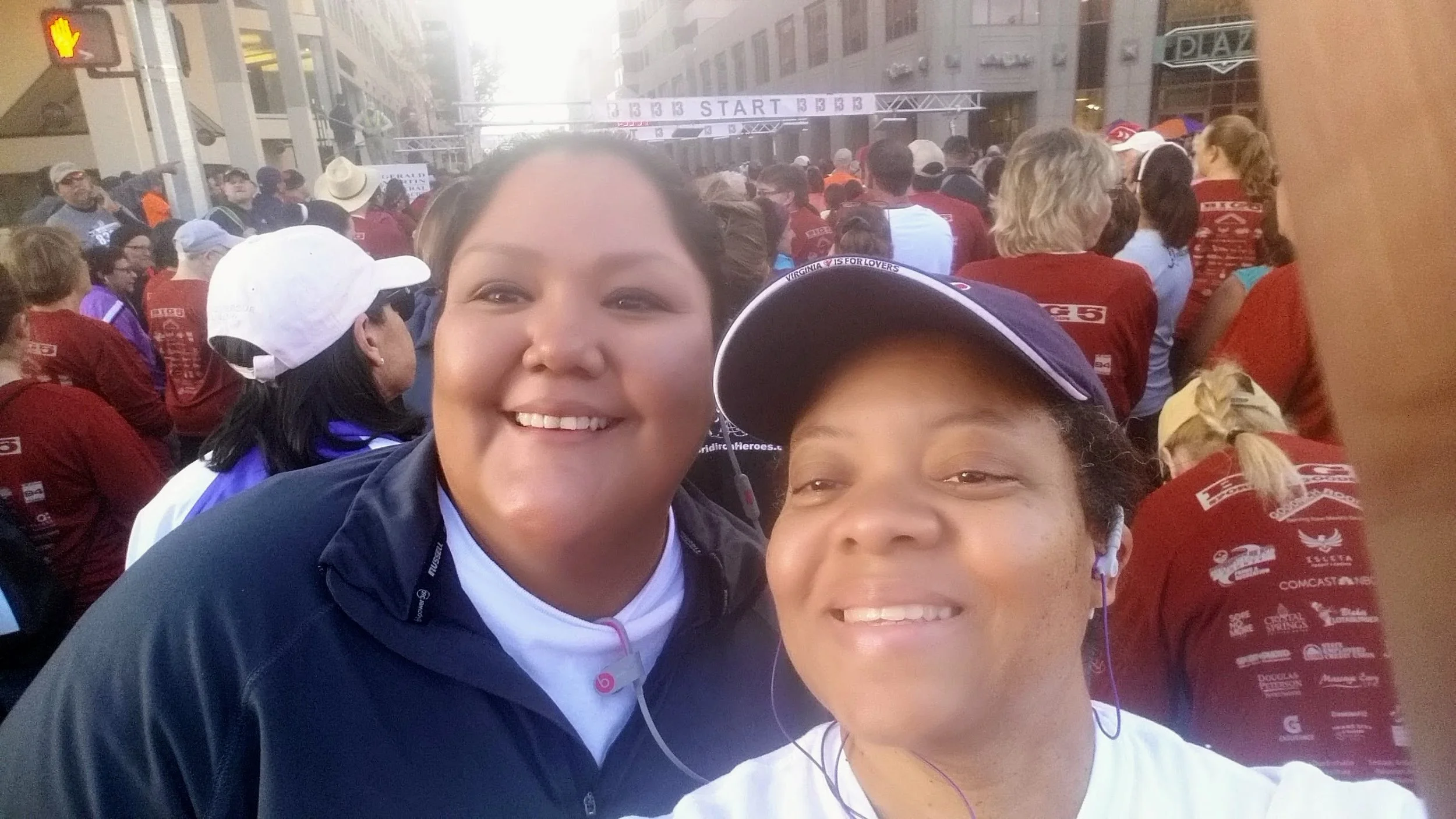 Two women taking a selfie at a large outdoor event, with a crowd of people in the background on a city street, near a marathon start line.