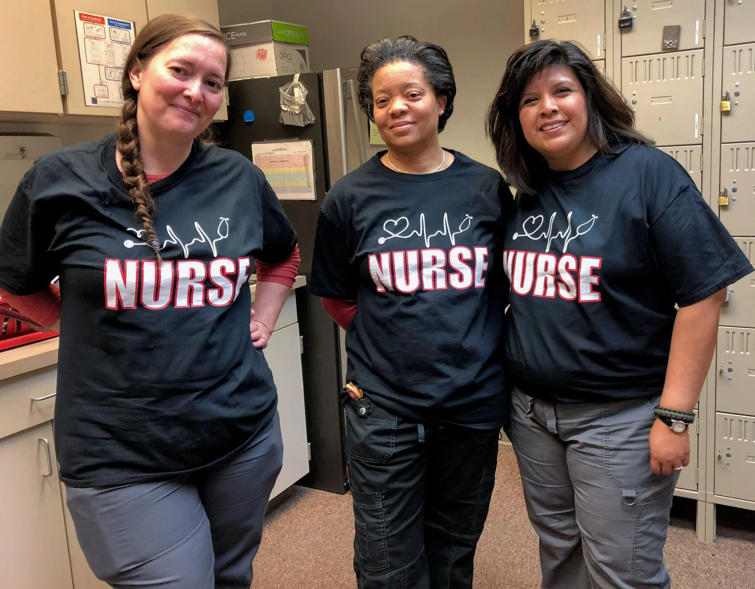 Three women wearing black nurse T-shirts standing in a room with lockers and cabinets.