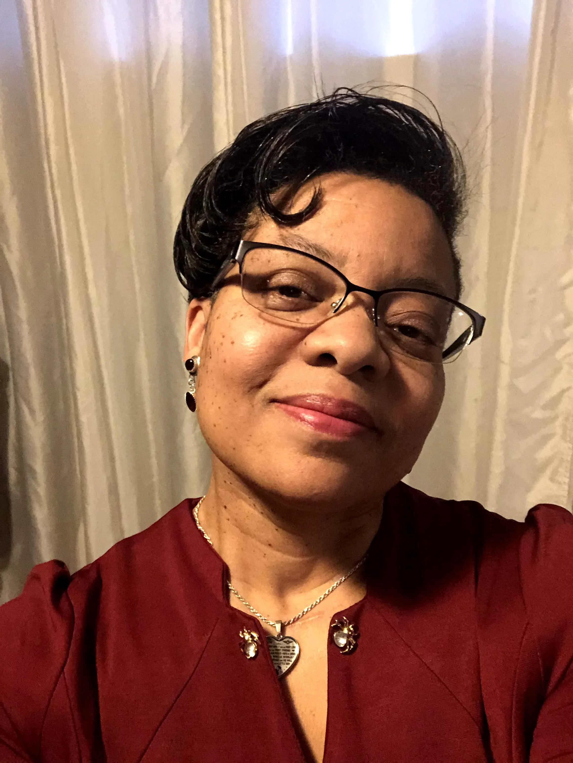 Close-up of a woman with short dark hair, wearing glasses, earrings, a necklace, a heart-shaped pendant, and a maroon blouse, standing in front of light-colored curtains.