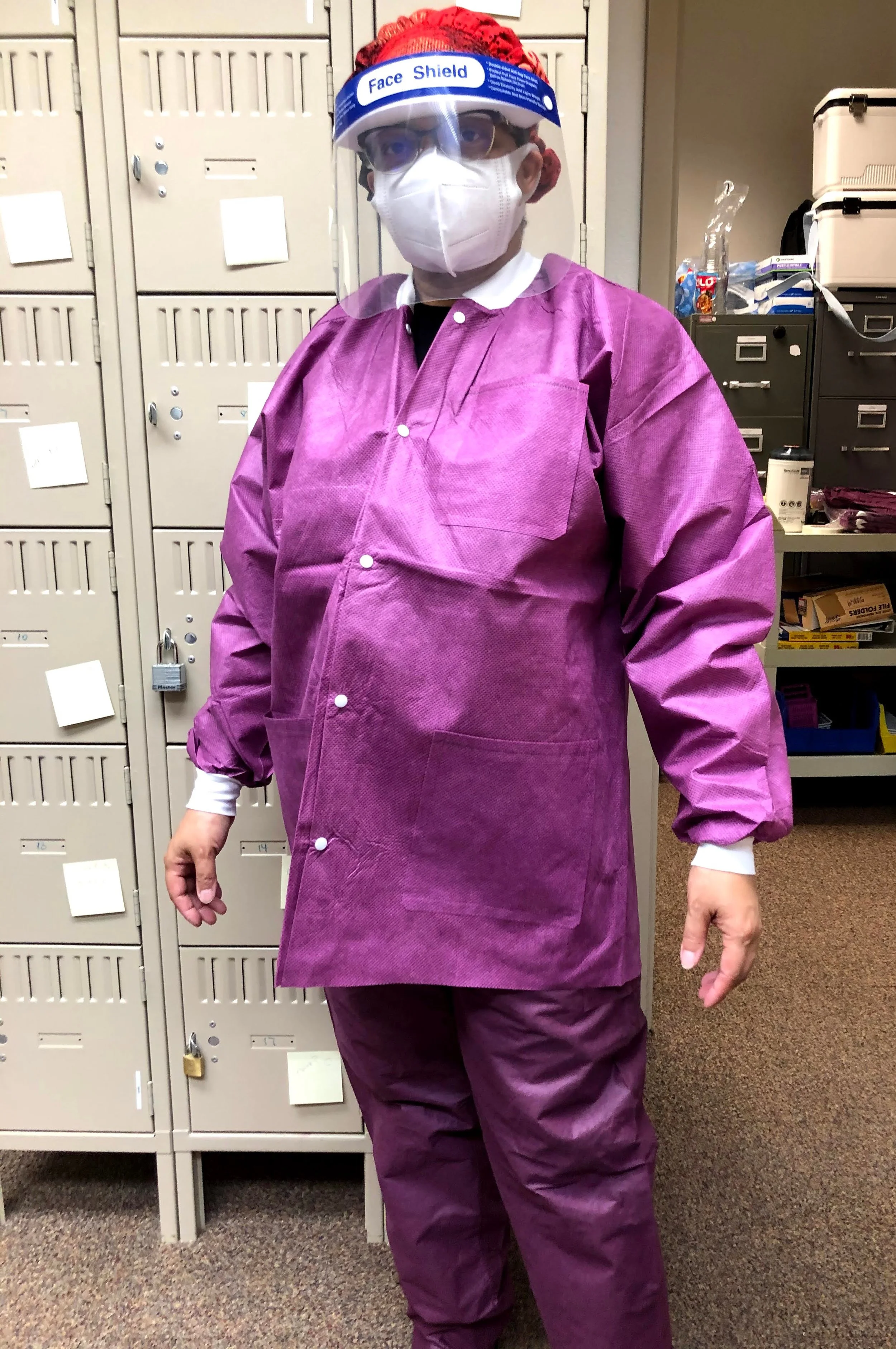 A person wearing purple protective clothing, a face mask, glasses, and a clear face shield standing in front of lockers in an indoor setting.