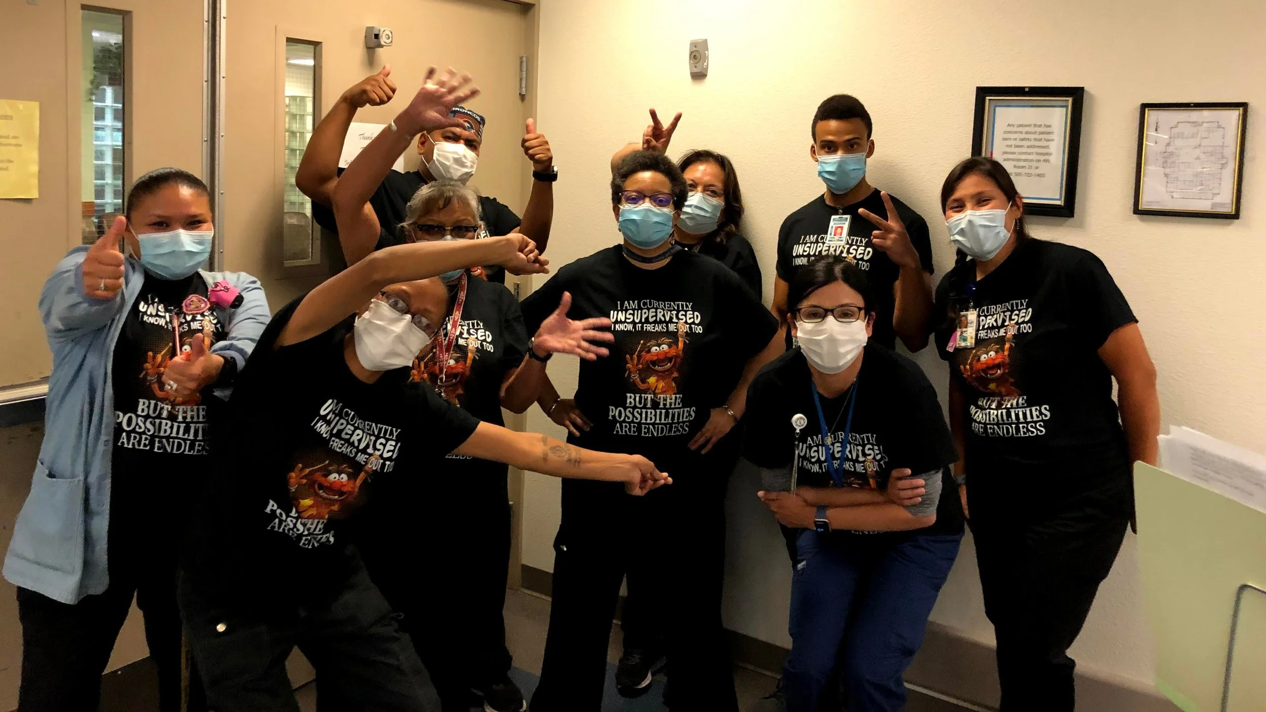 A diverse group of nine healthcare workers, all wearing masks and black T-shirts with a Sesame Street character and text, posing together in a hospital hallway. They are making various hand gestures and smiling.
