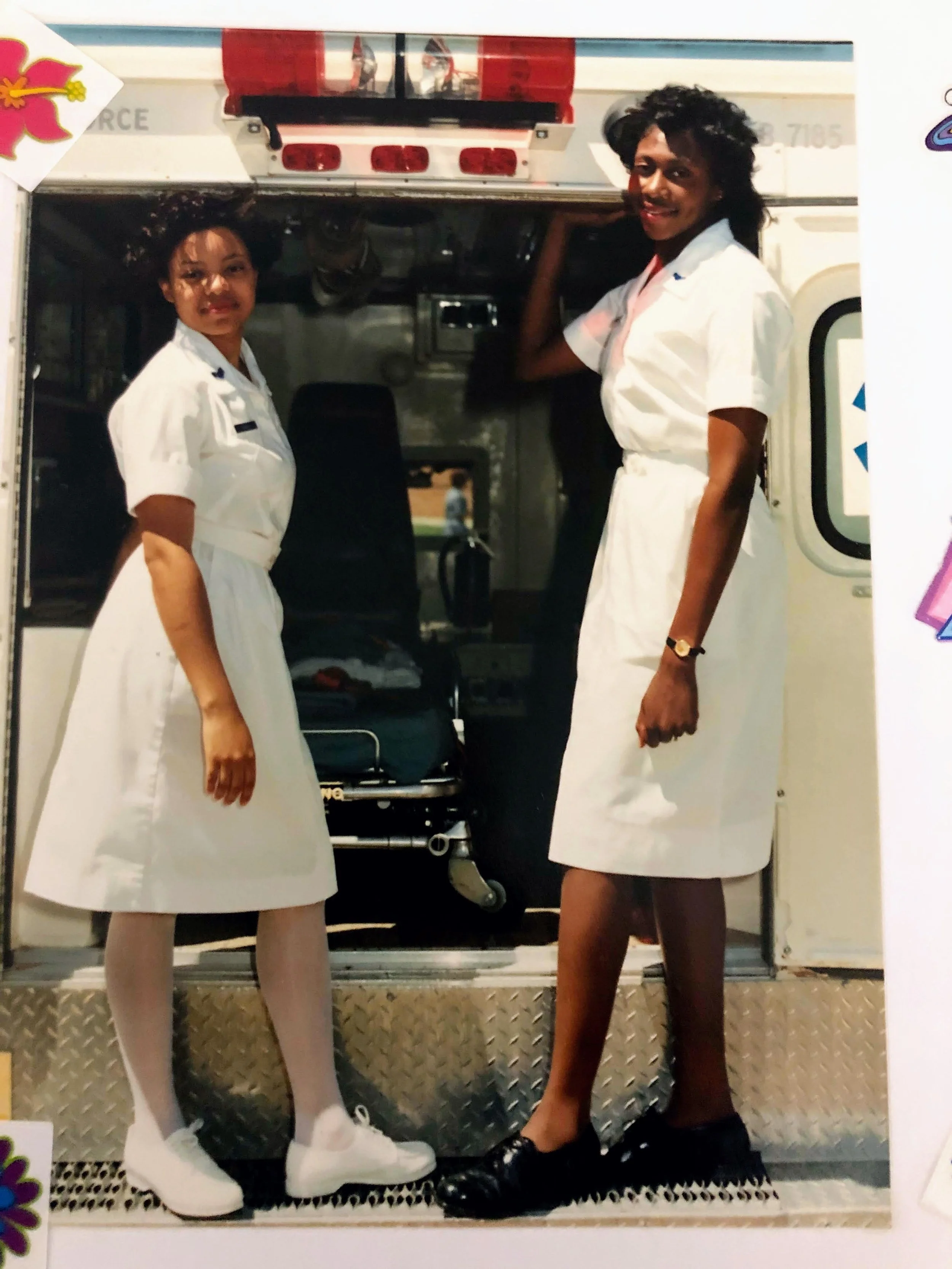 Two women dressed in nurse uniforms standing in front of an ambulance, smiling at the camera.