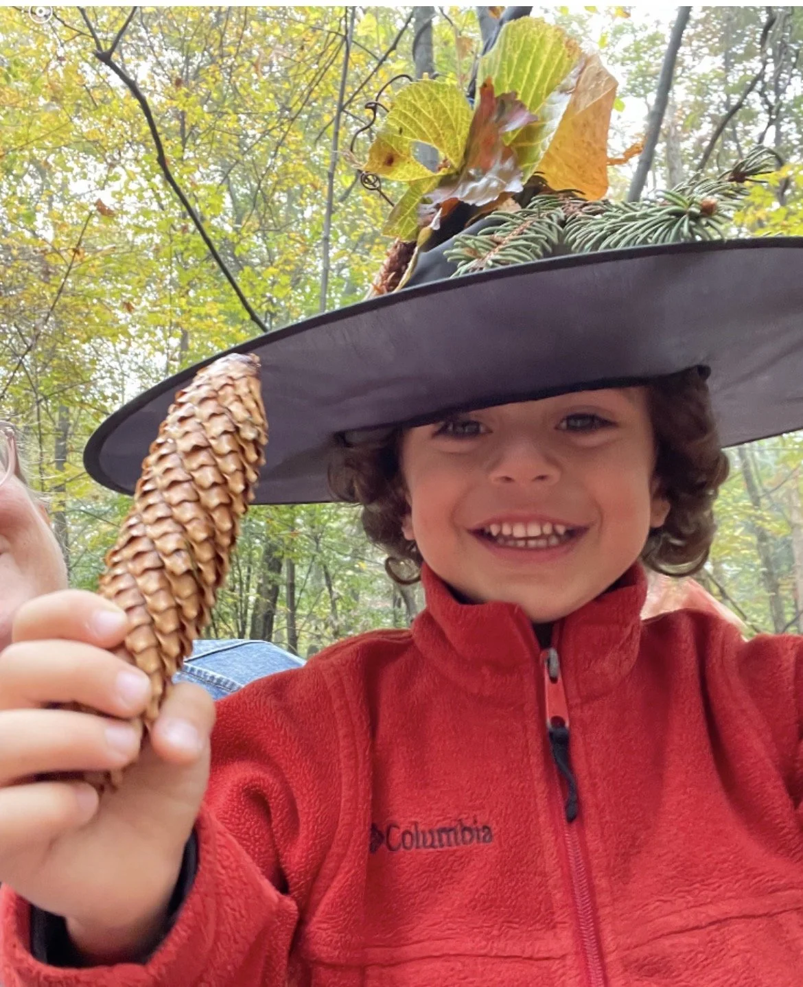 Smiling child in a witch hat decorated with leaves and pine, holding up a pinecone in the forest