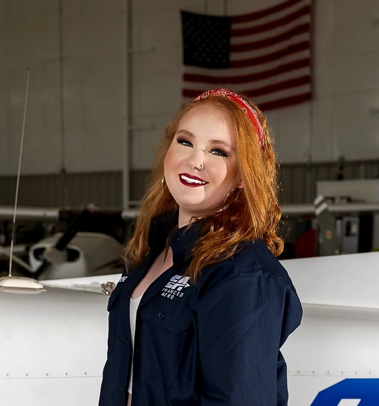 Smiling woman with red hair and a nose ring, wearing a blue jacket, standing in front of a white airplane inside a hangar with an American flag in the background.