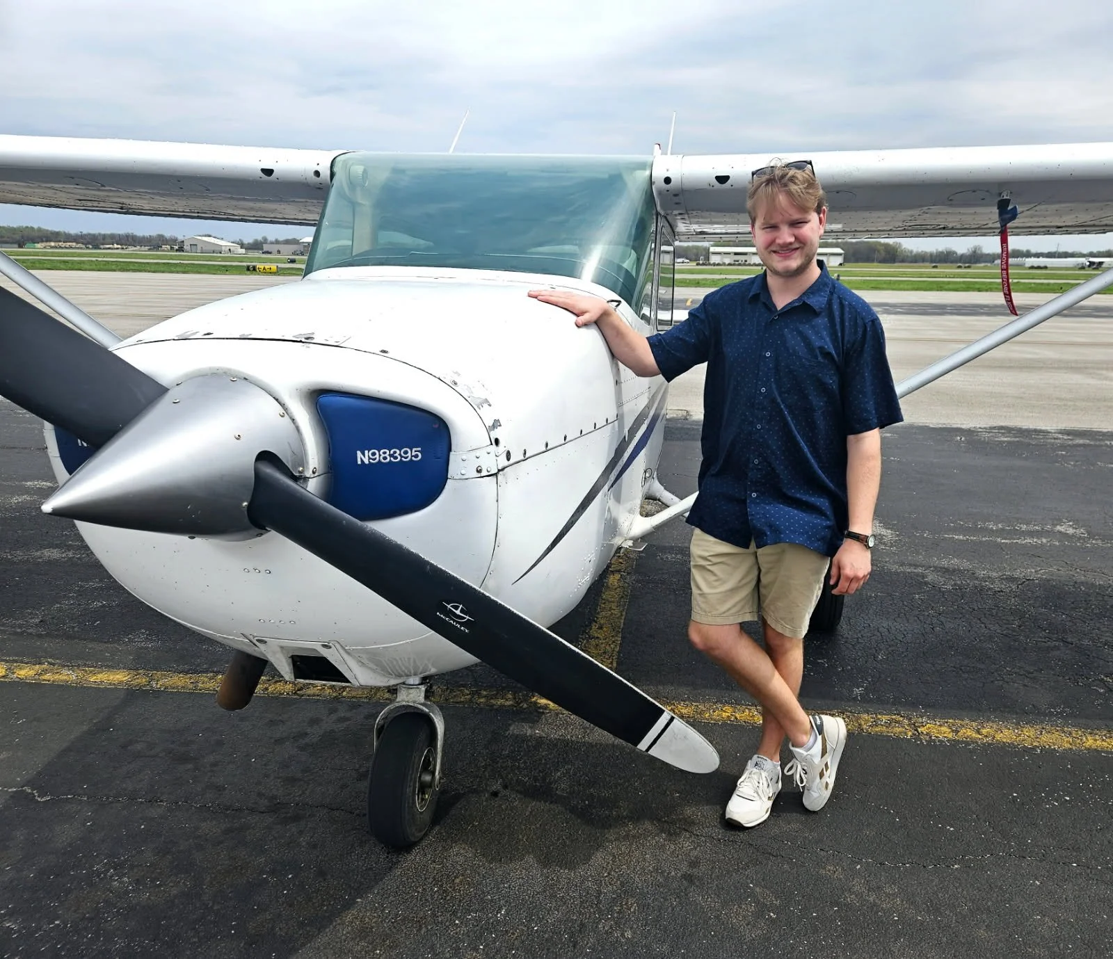 A young man in casual attire, wearing shorts and sneakers, standing next to a small white propeller airplane on an airport tarmac, with one hand resting on the plane.