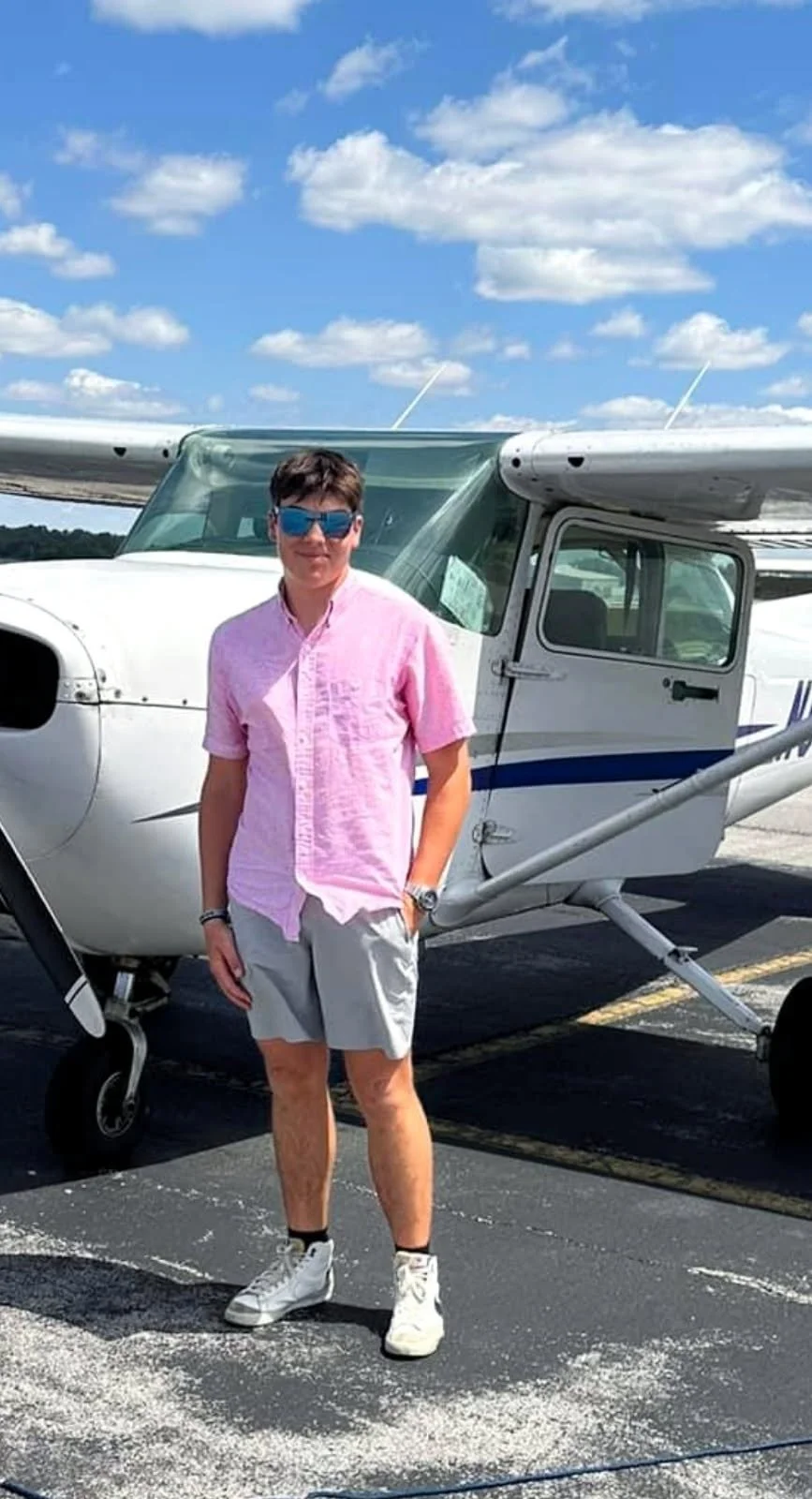 Person standing in front of a small airplane on an airport tarmac, wearing a pink shirt, gray shorts, white sneakers, and sunglasses, with a partly cloudy sky in the background.