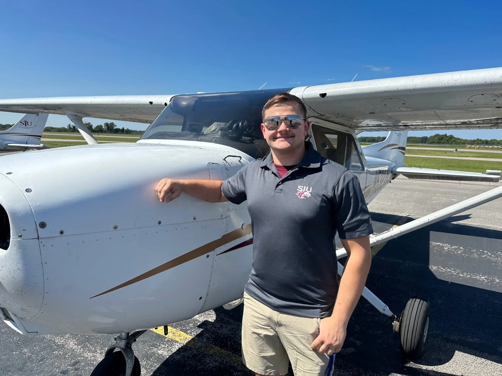 A young man wearing sunglasses and a gray polo shirt with a school logo, standing next to a small white airplane on a sunny day at an airport.