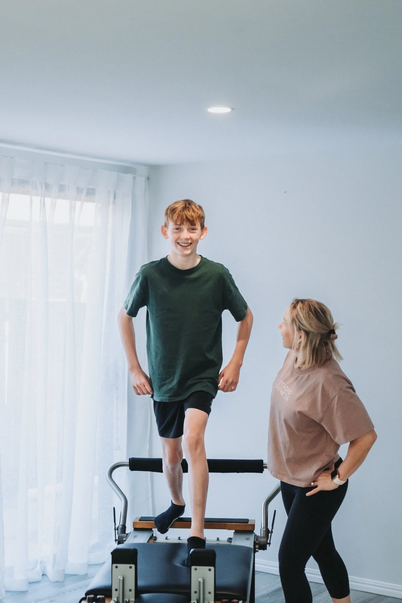 Mel standing next to her son on the reformer; her son beaming in a huge smile as he works the exercise through