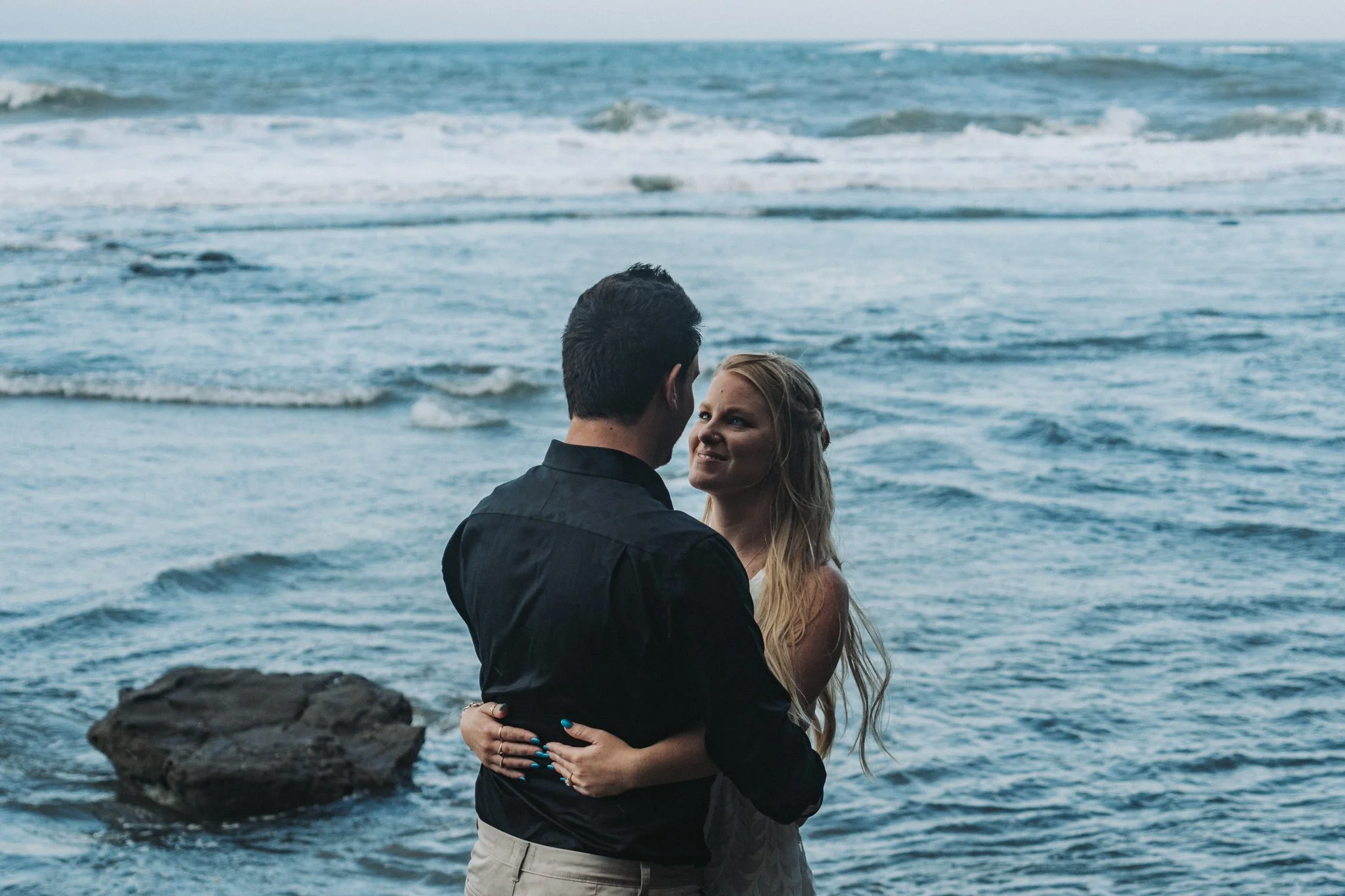 Young couple facing each other hugging, brides face in view with so much love lighting up her eyes as she gazes at her husband. While the afternoon's soft light bounds of the waves rolling in behind them at Shelly Beach, QLD.