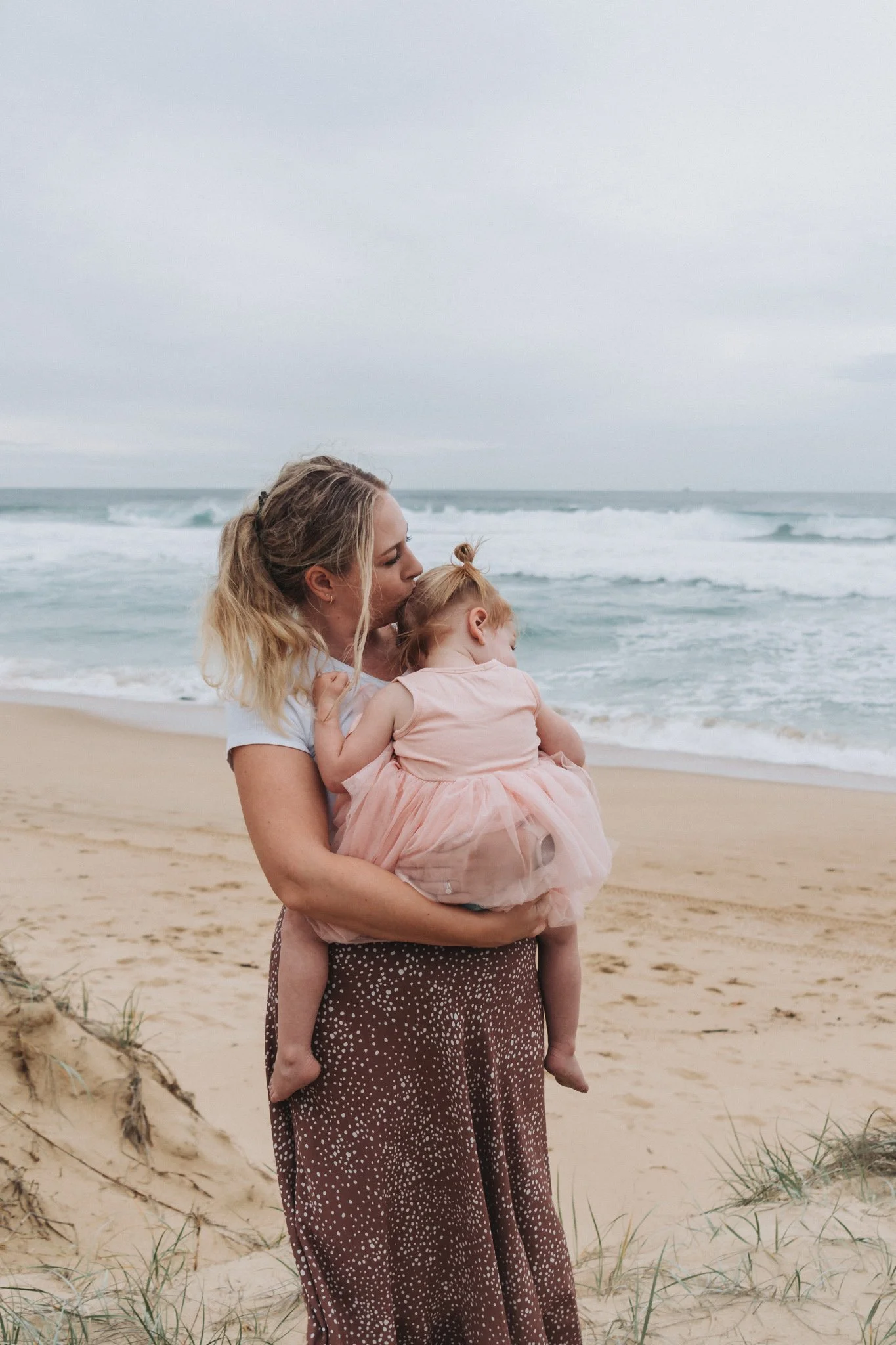 Mum holding her little girl, kissing her head while the little girl snuggly looks out to sea