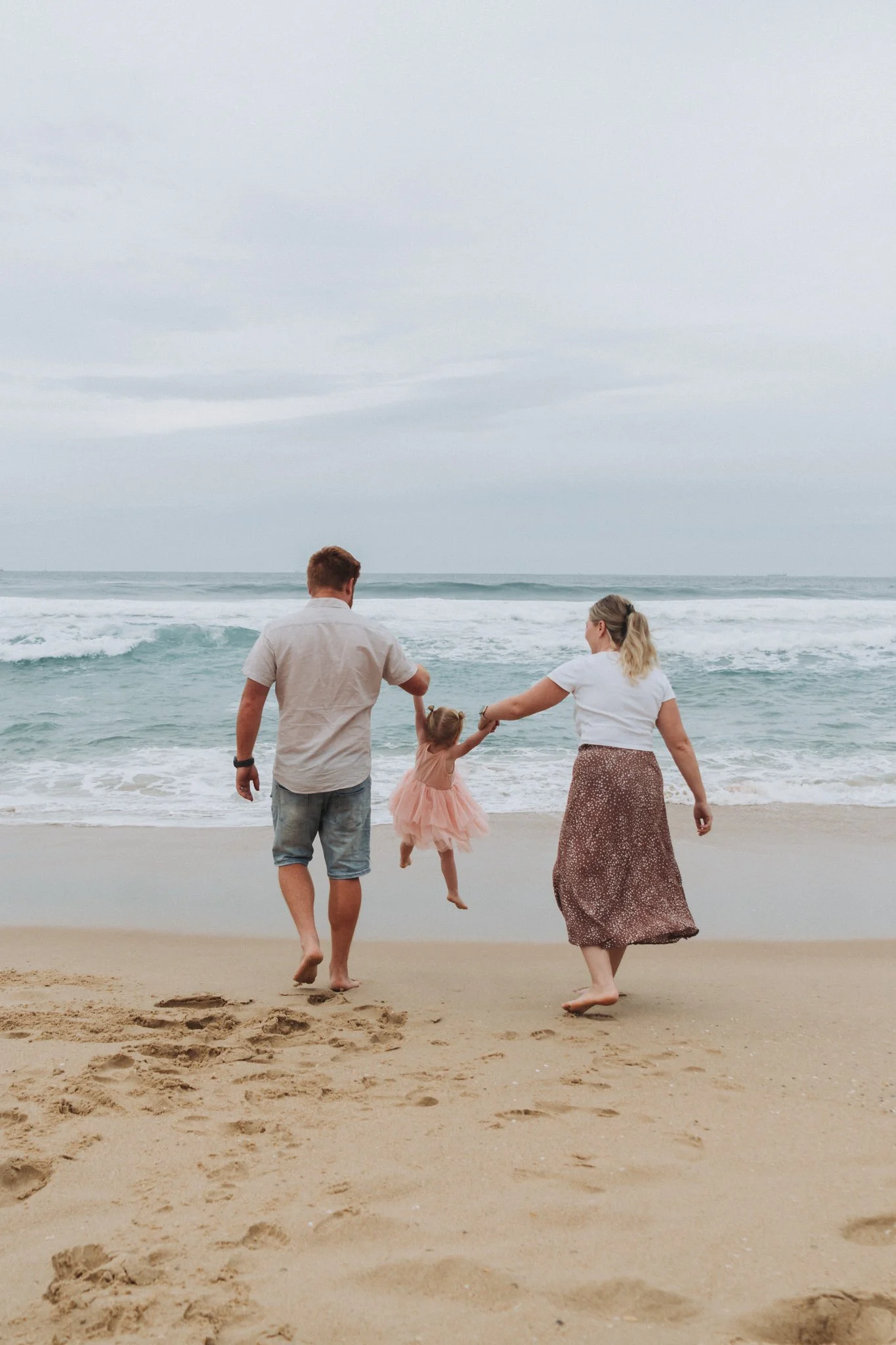 Young family on three on Kawana Beach Sunshine Coast. Holding hand, Mum and Dad swing their little girl between them