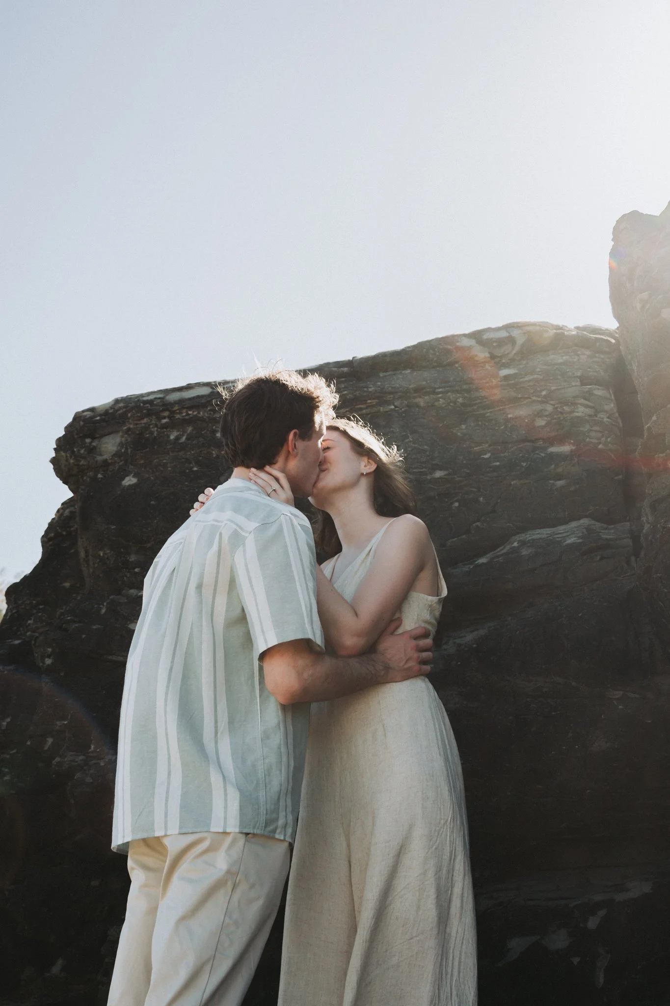 Sweet kisses between young , freshly engaged couple below the cliffs of Point Cartwright, enveloped in soft, warm light flares