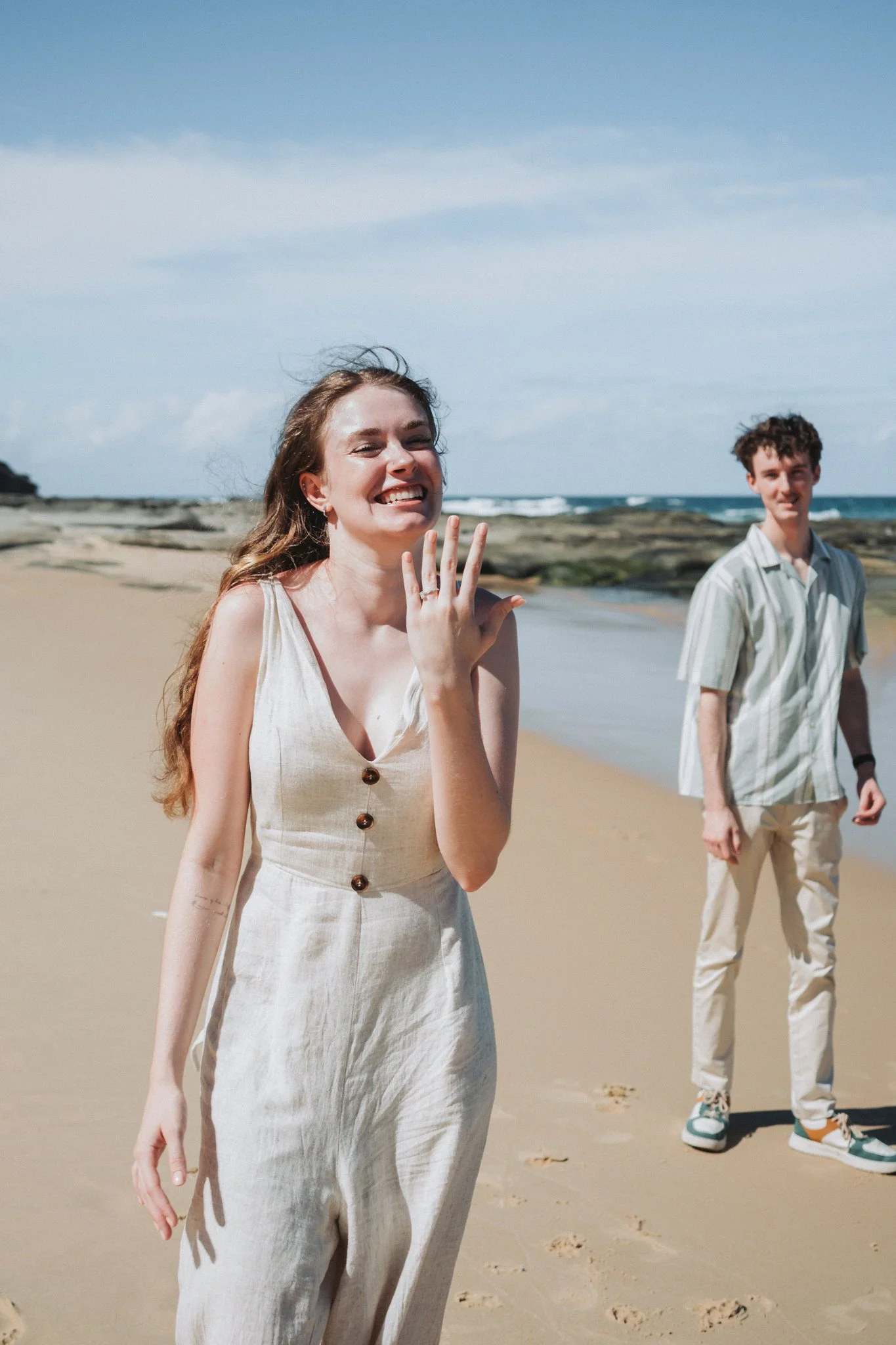 Young, freshly engaged couple celebrating on Sunshine Coast's iconic beach at Point Cartwright. Radiant bride to be laughing and running to the camera showing off her stunning engagement ring
