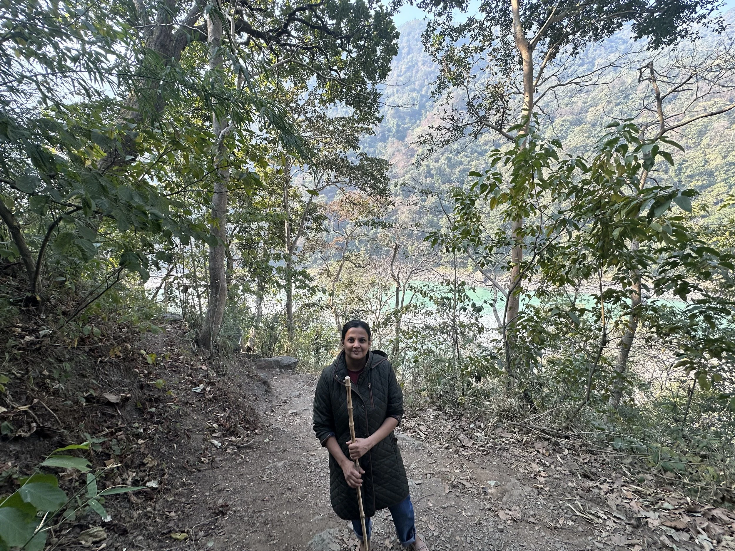 A woman standing next to a trail in a forested area with mountains and trees in the background, holding a walking stick.