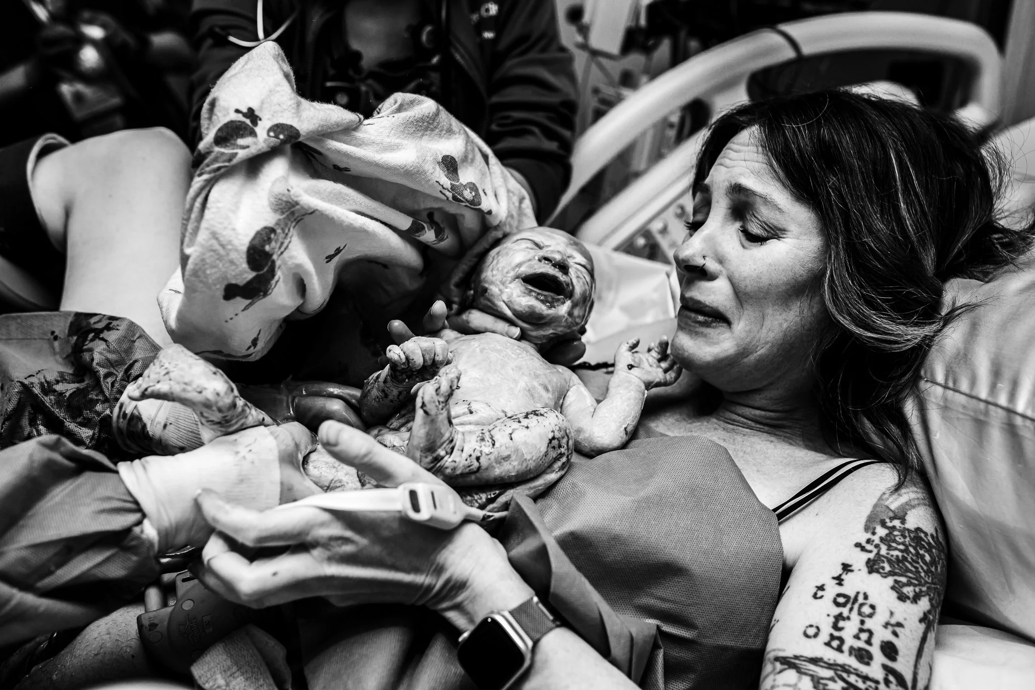 A woman lying in a hospital bed immediately after delivering a baby, holding her newborn who is crying. Medical staff is assisting with the delivery.