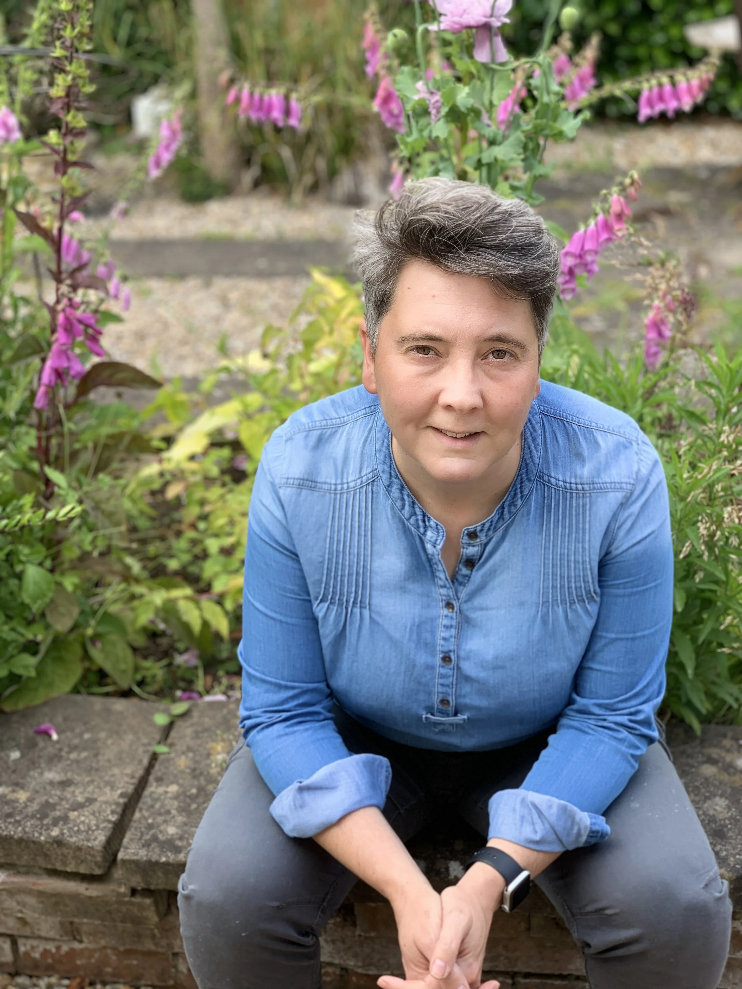 A person with short, gray and dark hair sits on a brick ledge in a garden with pink and purple flowers and green foliage.