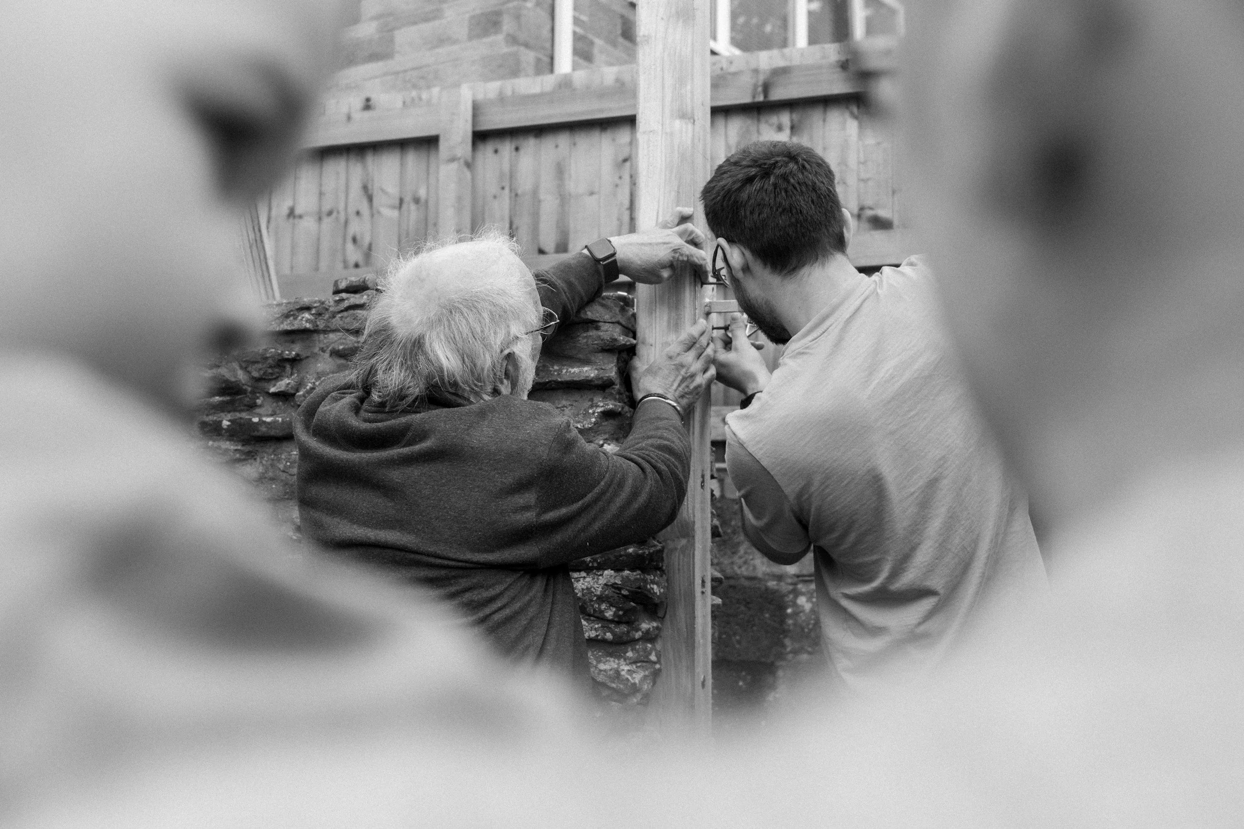 Two men working together on a woodworking project outdoors, with a wooden fence in the background, as seen through a blurred foreground frame.