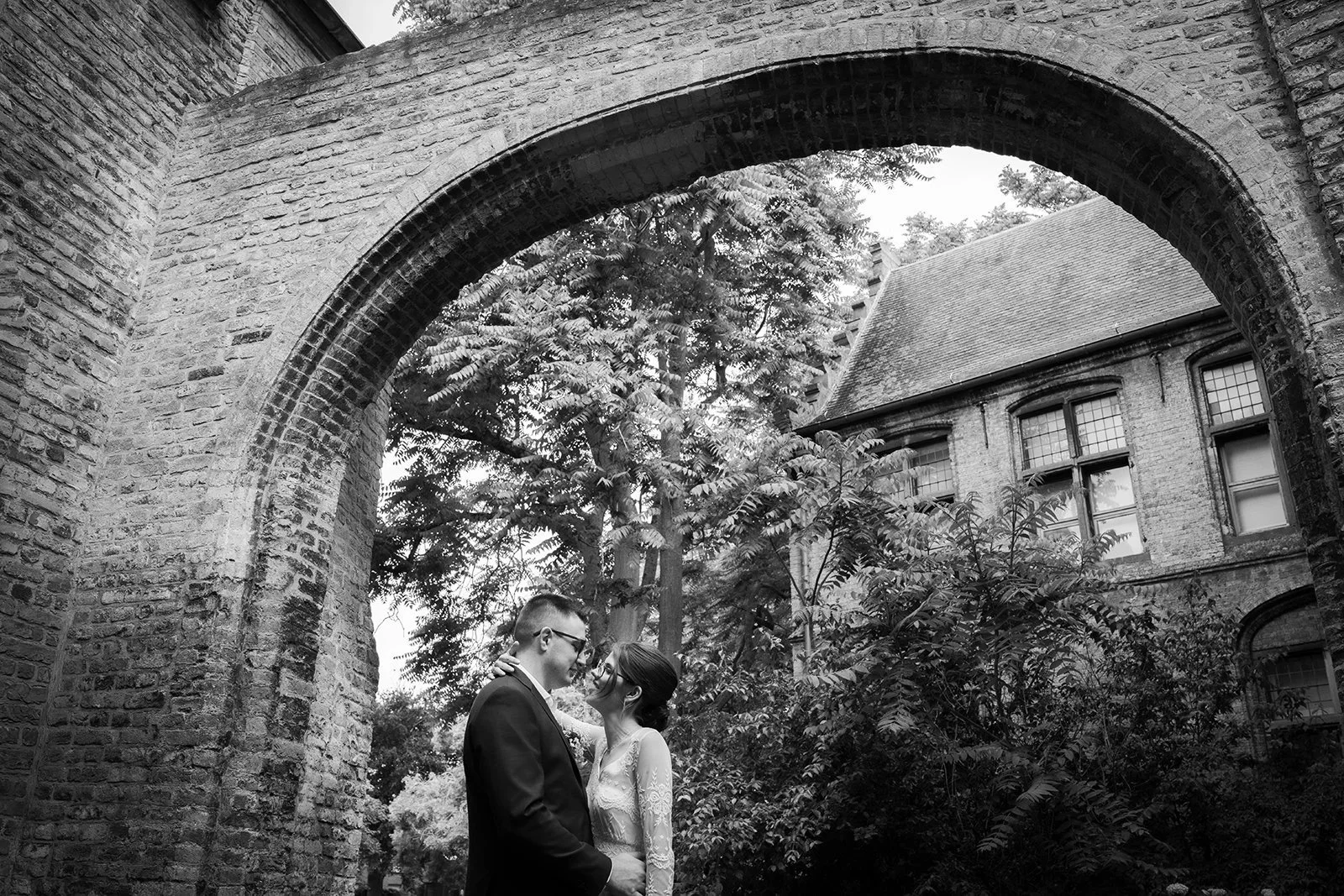 Een zwart-wit foto van een koppel op hun bruiloft, staande onder een oude stenen boog, omringd door bomen en historische gebouwen.