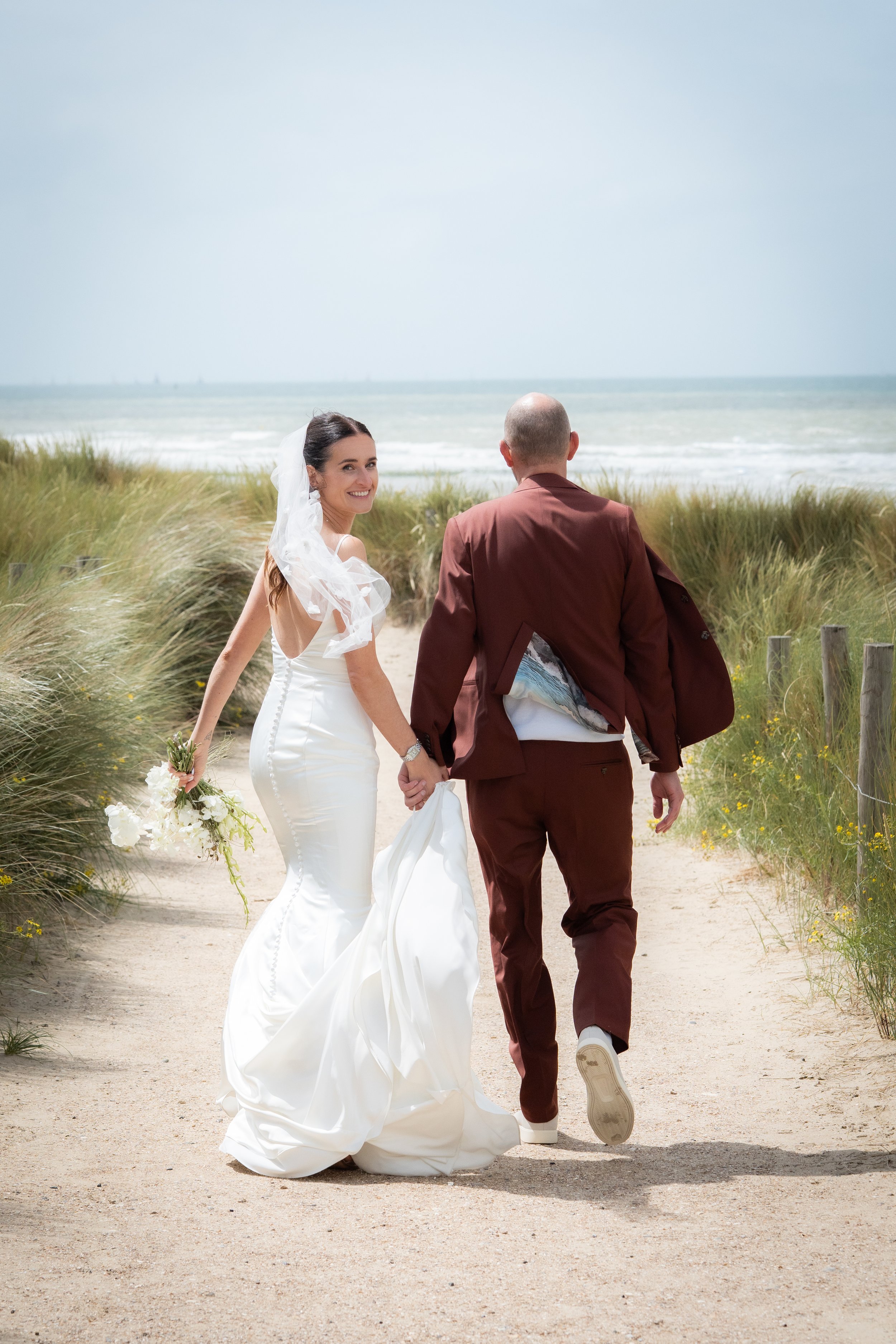 Een bruid en bruidegom lopen hand in hand op een zandpad langs het strand, omgeven door duinen en gras, met de oceaan op de achtergrond.