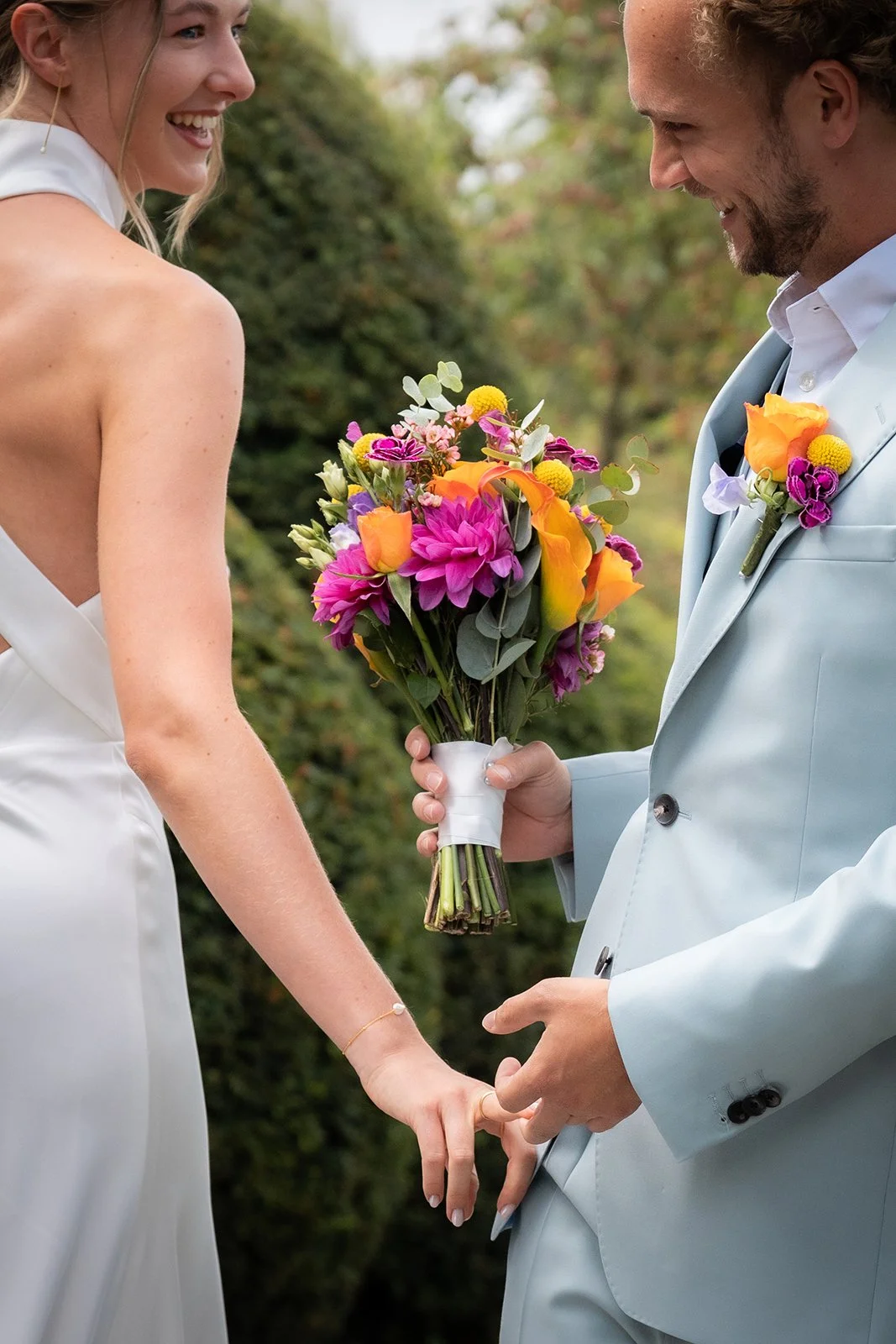 Een stel op hun bruiloft, hand in hand, met de bruid die een kleurrijke bloemenbundel vasthoudt, buiten met groenigend landschap.
