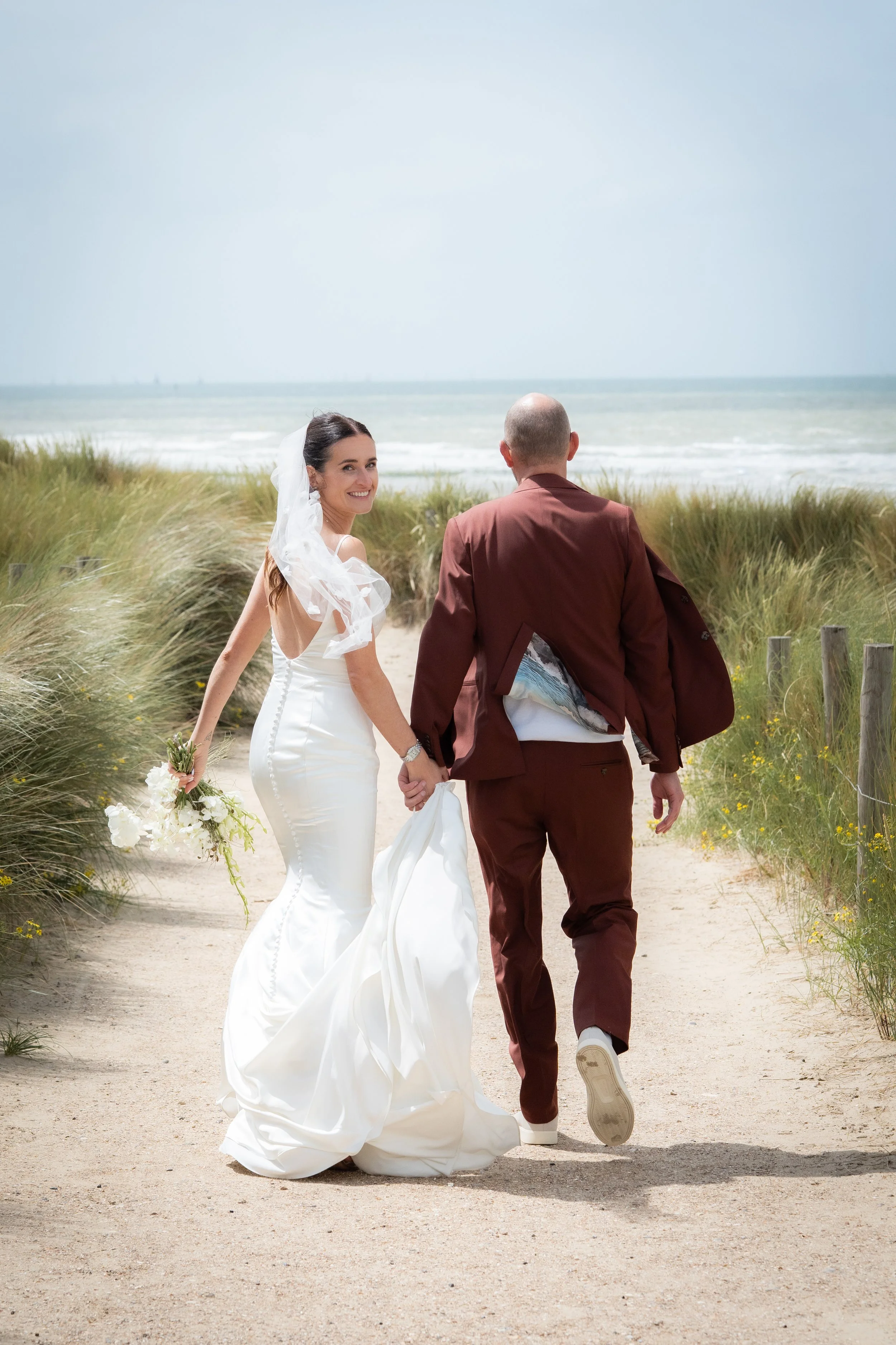 Een koppel dat hand in hand wandelt op een strand, de vrouw in een witte trouwjurk en de man in een bruin pak, met de oceaan op de achtergrond.