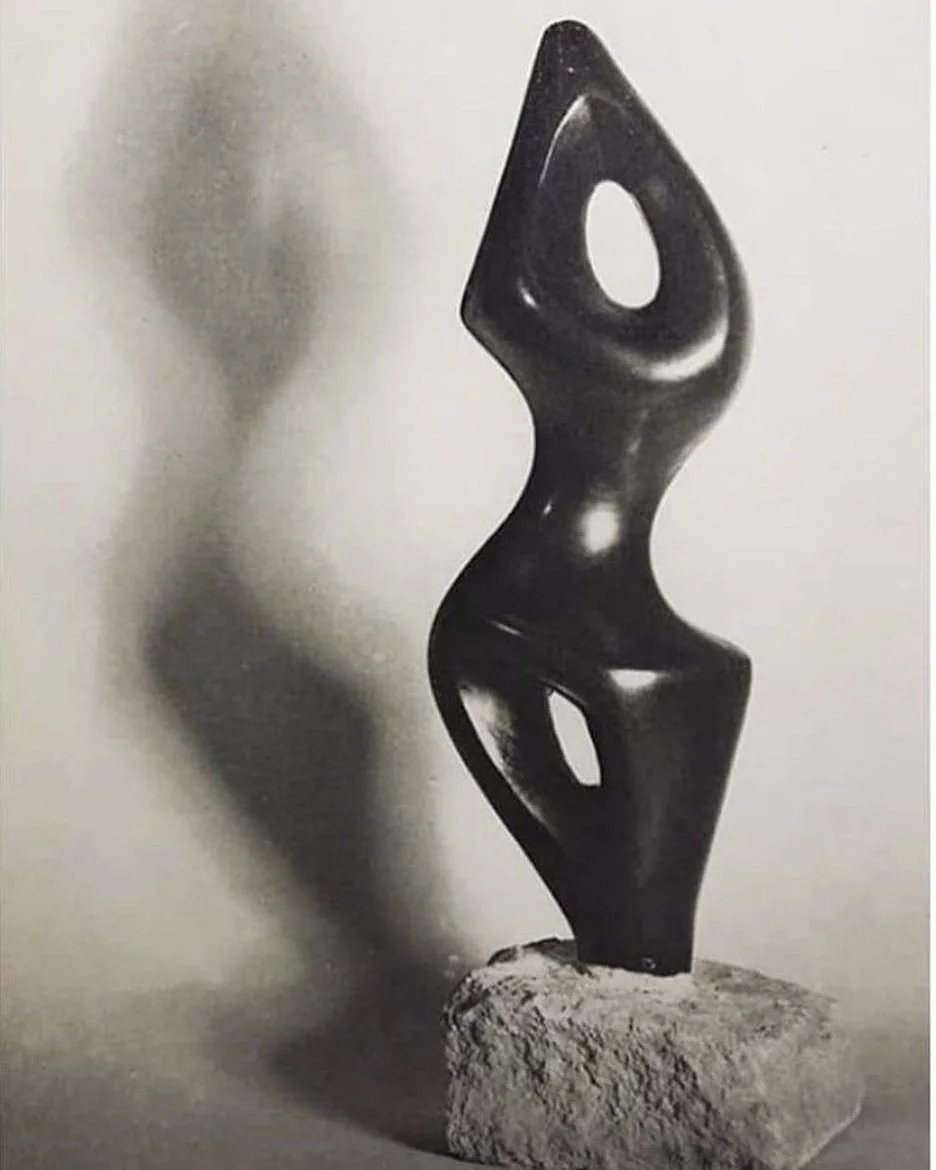 Abstract black sculpture on a stone base with smooth, curved shapes and hollow sections, creating a shadow on the wall.
