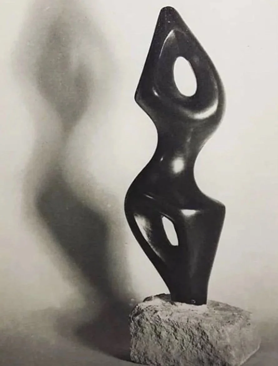 Abstract black sculpture on a stone base with smooth, curved shapes and hollow sections, creating a shadow on the wall.