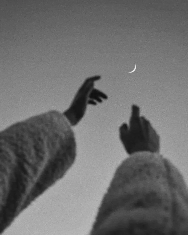 Black and white image of two hands reaching towards a crescent moon in the sky.