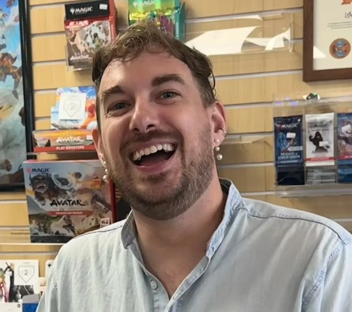 A smiling man with light brown hair, beard, and earrings, wearing a light-colored shirt, standing in front of a display of Magic: The Gathering cards in a store.