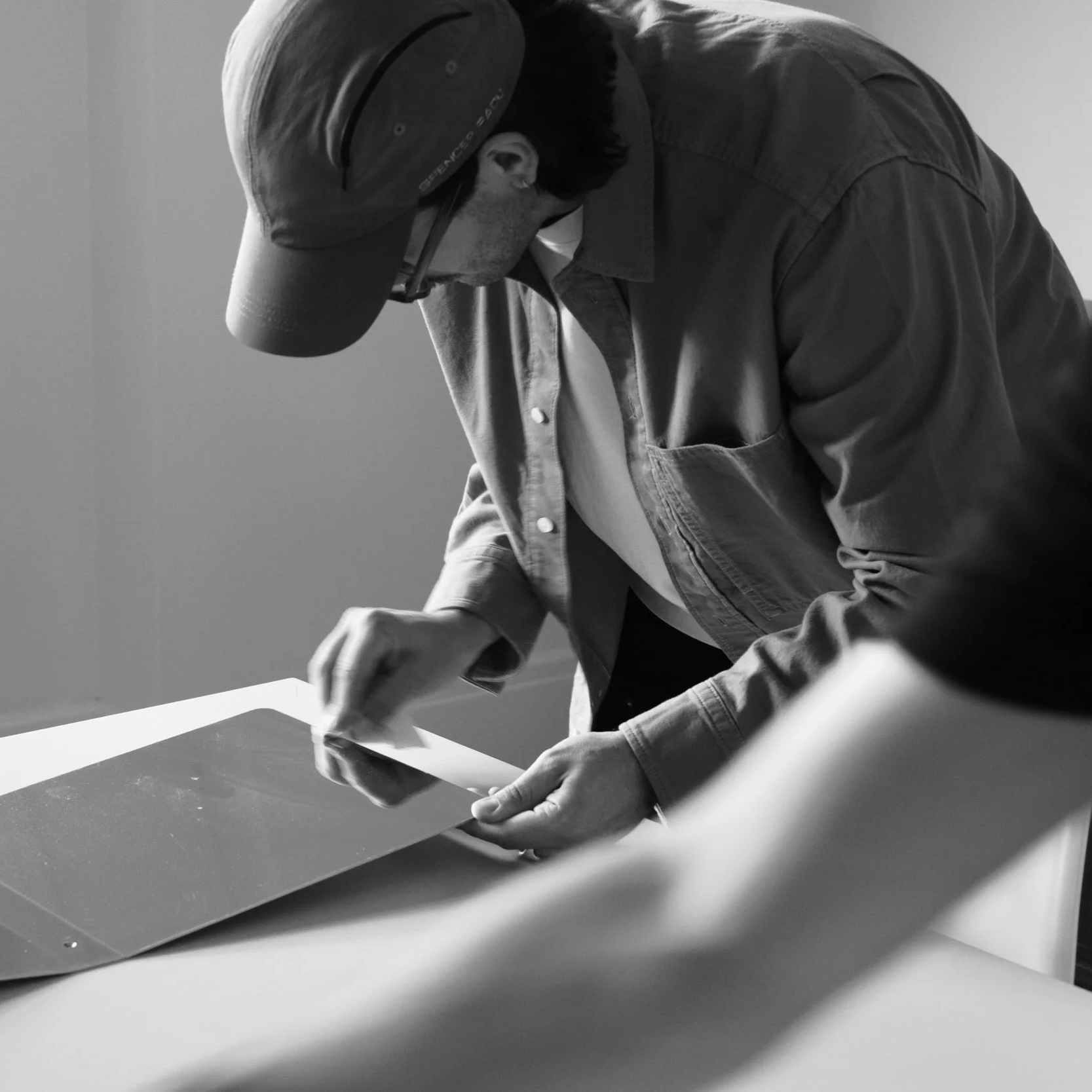 Gilbert Fortin wearing a cap and glasses is inspecting or working on a rectangular object on a table in a room with plain walls.