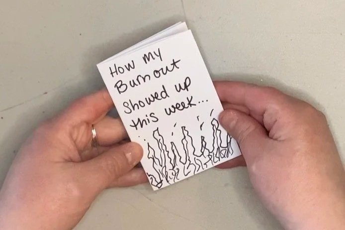 Two hands holding a small handmade zine with the handwritten title "How my Burnout Showed Up This Week" and a simple doodle of flame-like figures along the bottom.