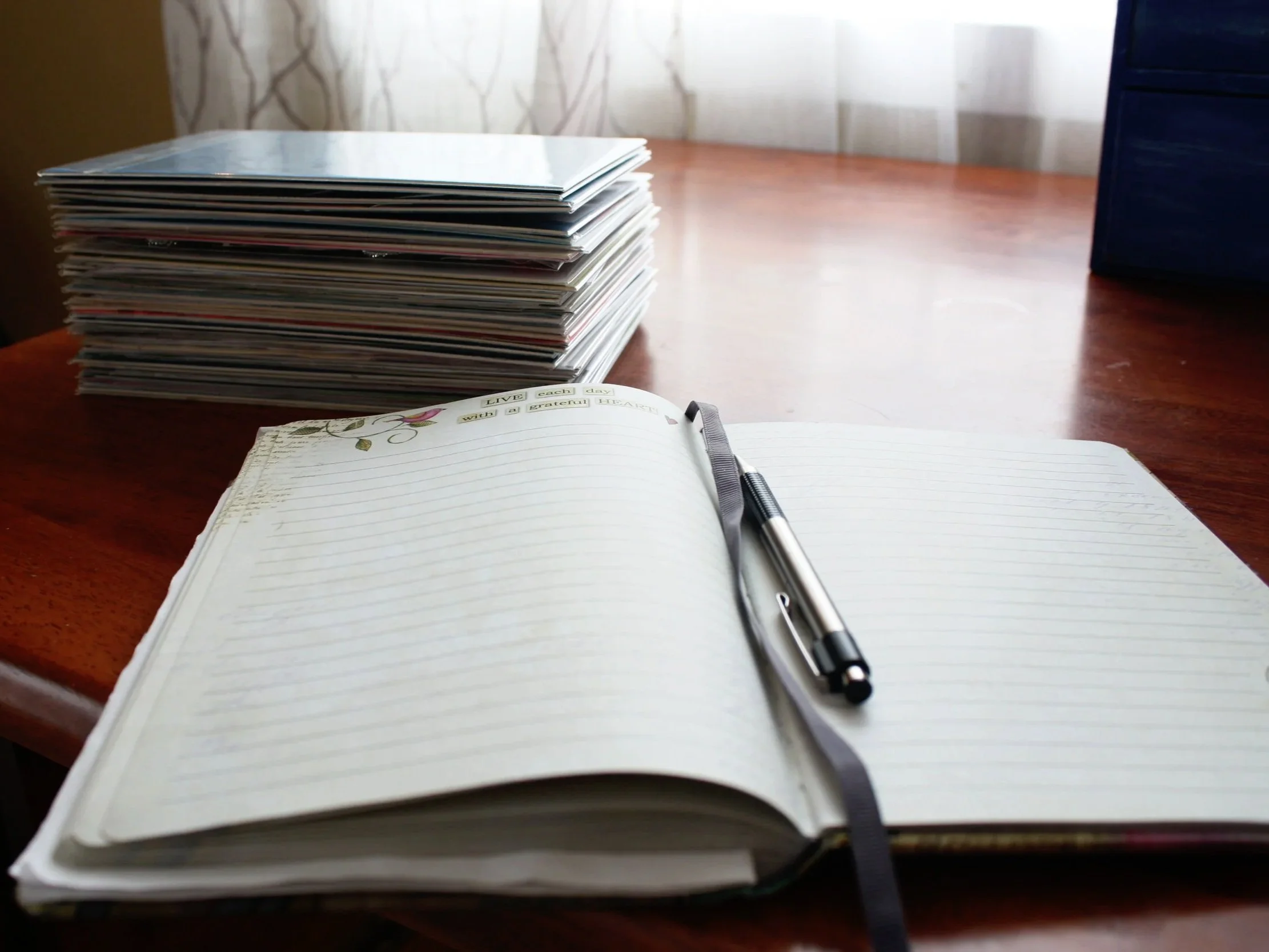 Open notebook with blank lined pages with a pen on top. Behind the notebook there is a large stack of SoulCollage® cards.