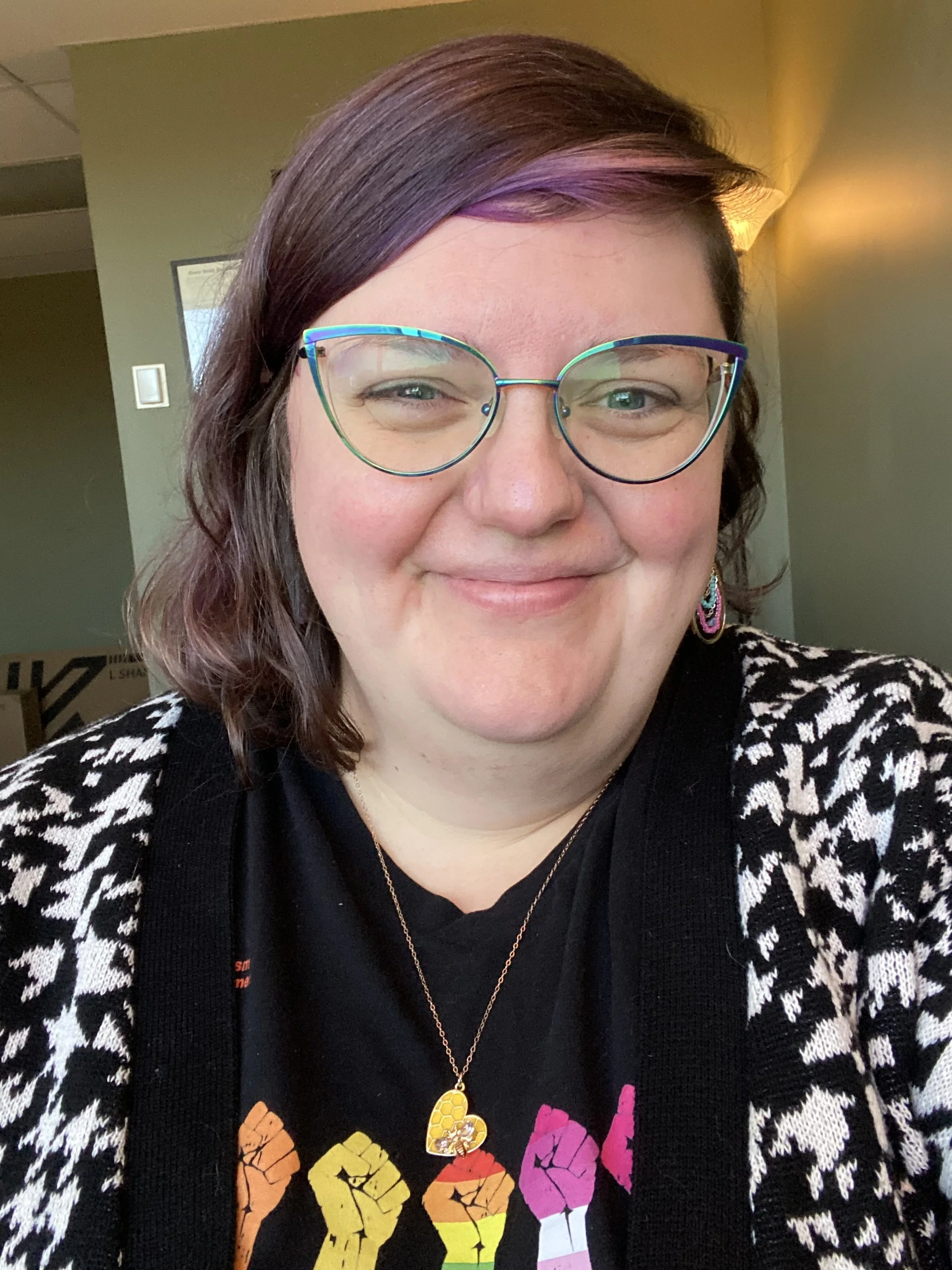 Close up of Rebel Wolfe, a white, plus size, femme person with glasses, brown and purple hair, smiling and wearing a pride t shirt and a black and white cardigan.