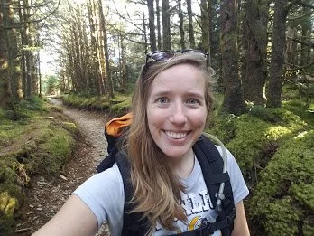 A woman smiling while hiking in a forest with trees on each side and sunlight streaming through.