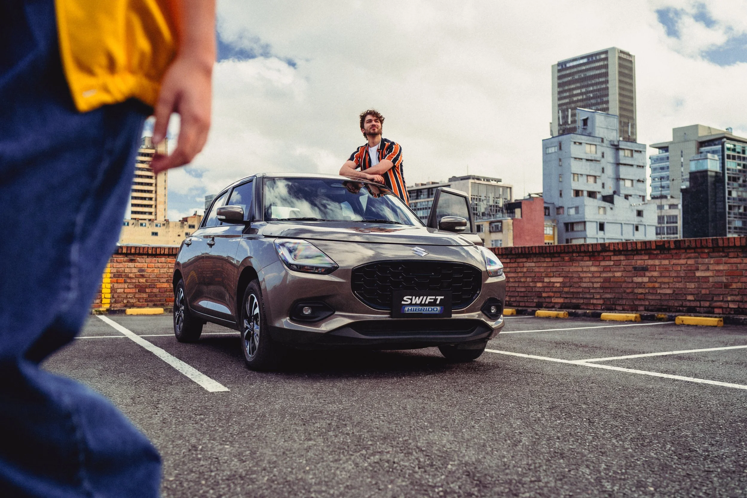 Hombre de pie junto a un coche compacto de color gris en un estacionamiento en la ciudad, con edificios altos y cielo nublado de fondo.