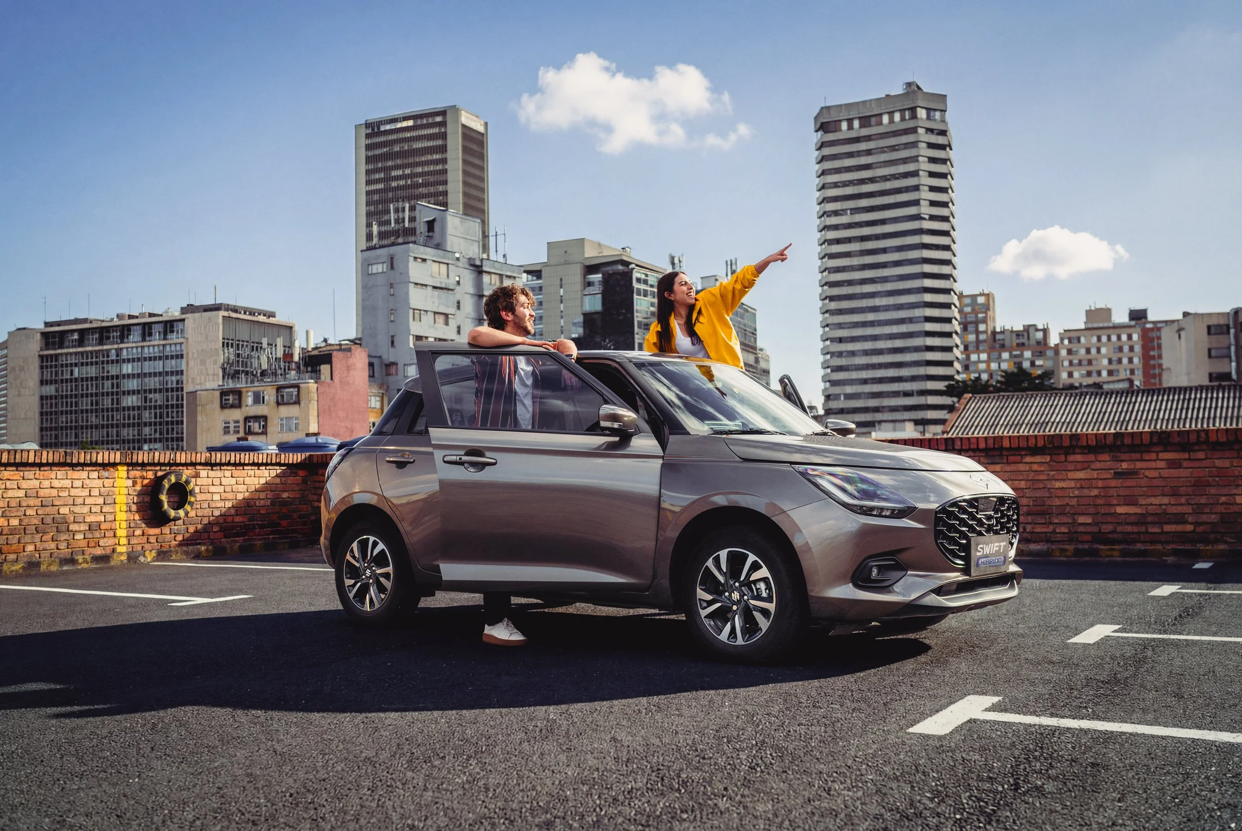 Dos personas disfrutan en un techo de estacionamiento urbano con edificios altos al fondo, uno de ellos mira hacia un lado y la otra apunta hacia adelante, ambas parecen estar felices.