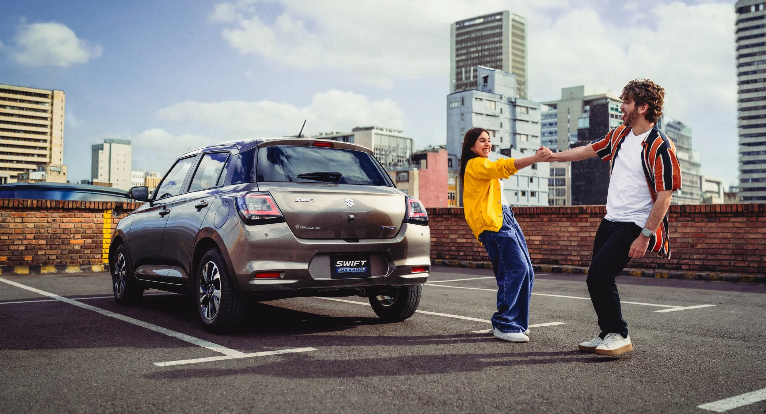 Una pareja feliz bailando y sujetándose de las manos en un estacionamiento con una ciudad de fondo y un coche plateado cercano.
