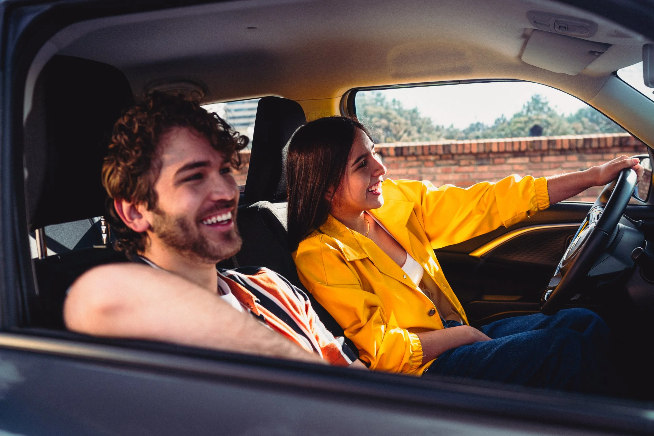 Dos jóvenes disfrutando en el coche, el hombre con cabello rizado y barba usa una camiseta de manga larga, la mujer con cabello liso y oscuro lleva una chamarra amarilla, ambos sonrientes.