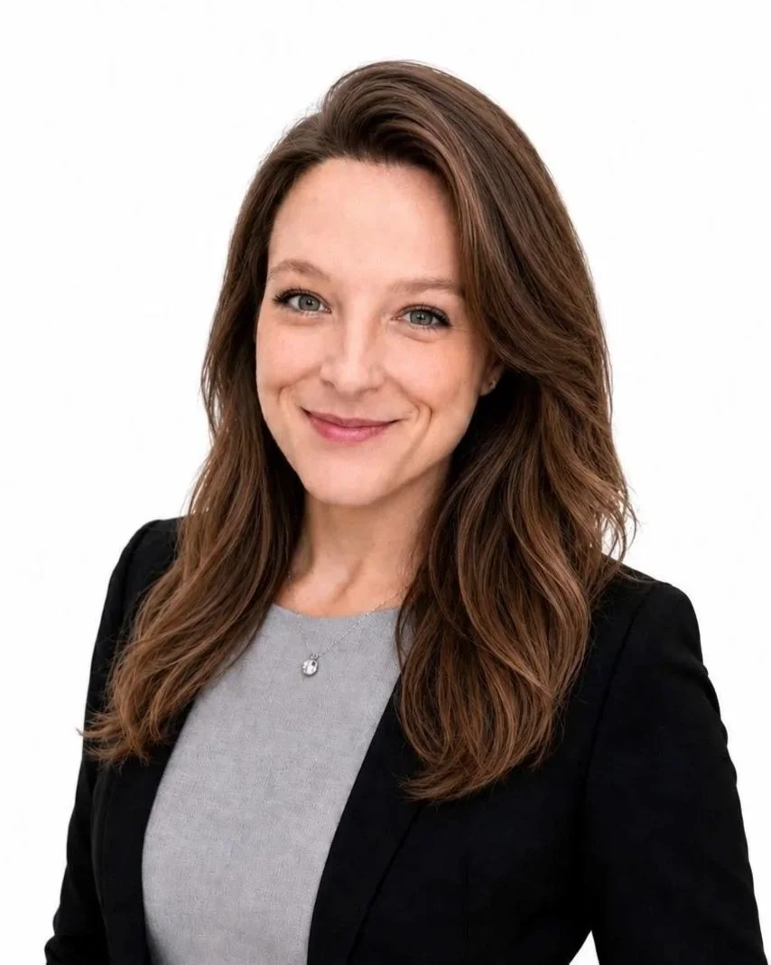Professional headshot of a smiling woman with long brown hair, wearing a black blazer and a light gray top, against a white background.