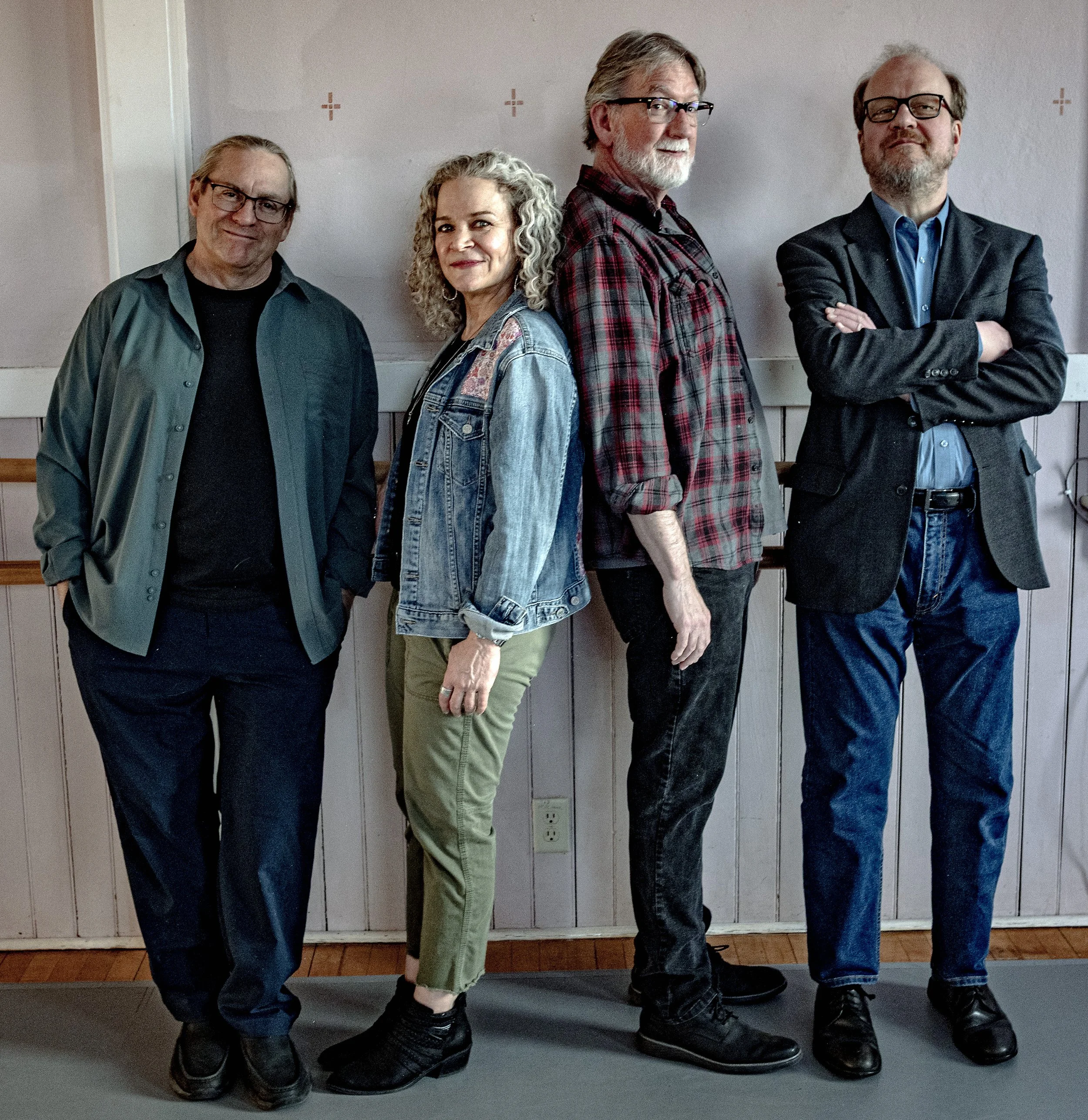 Group of four adults standing in a line against a plain wall, posing for the photo with relaxed expressions and one with crossed arms.