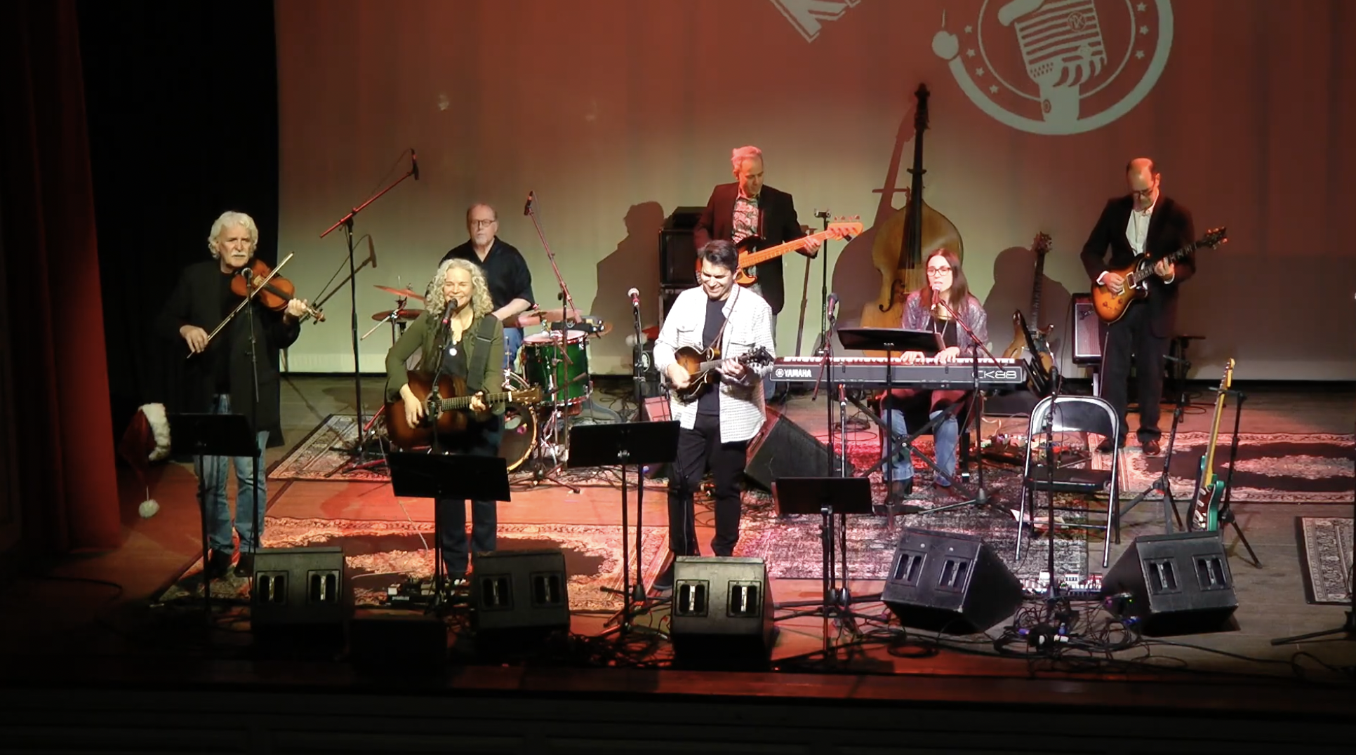 A group of seven musicians performing on stage with various instruments including a violin, guitars, keyboard, drums, an upright bass, and a microphone. The stage has a red wall with a logo in the background, and several rugs and musical equipment ar