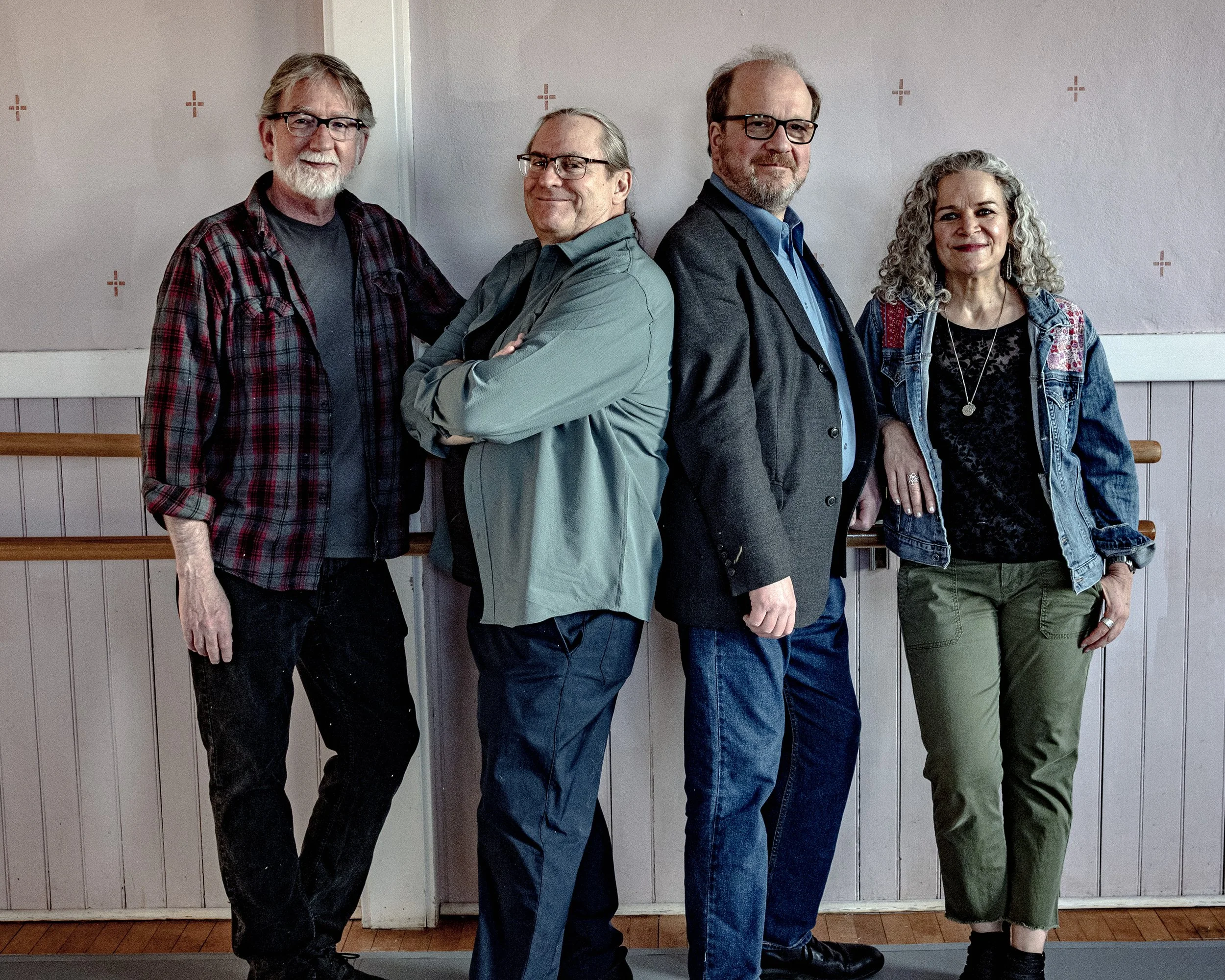 Four adults standing side by side indoors, posing for a photo, against a plain wall with a wooden railing in the background. The group includes three men and one woman, all dressed casually and smiling.