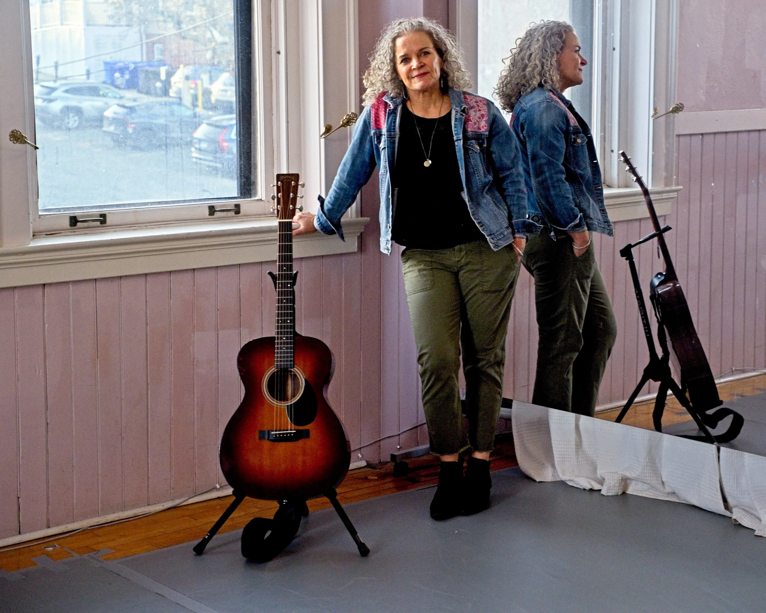 A woman with curly gray hair in a denim jacket and green pants standing by a window, with an acoustic guitar resting on a stand beside her, in a room with wood flooring and pinkish-paneled walls.