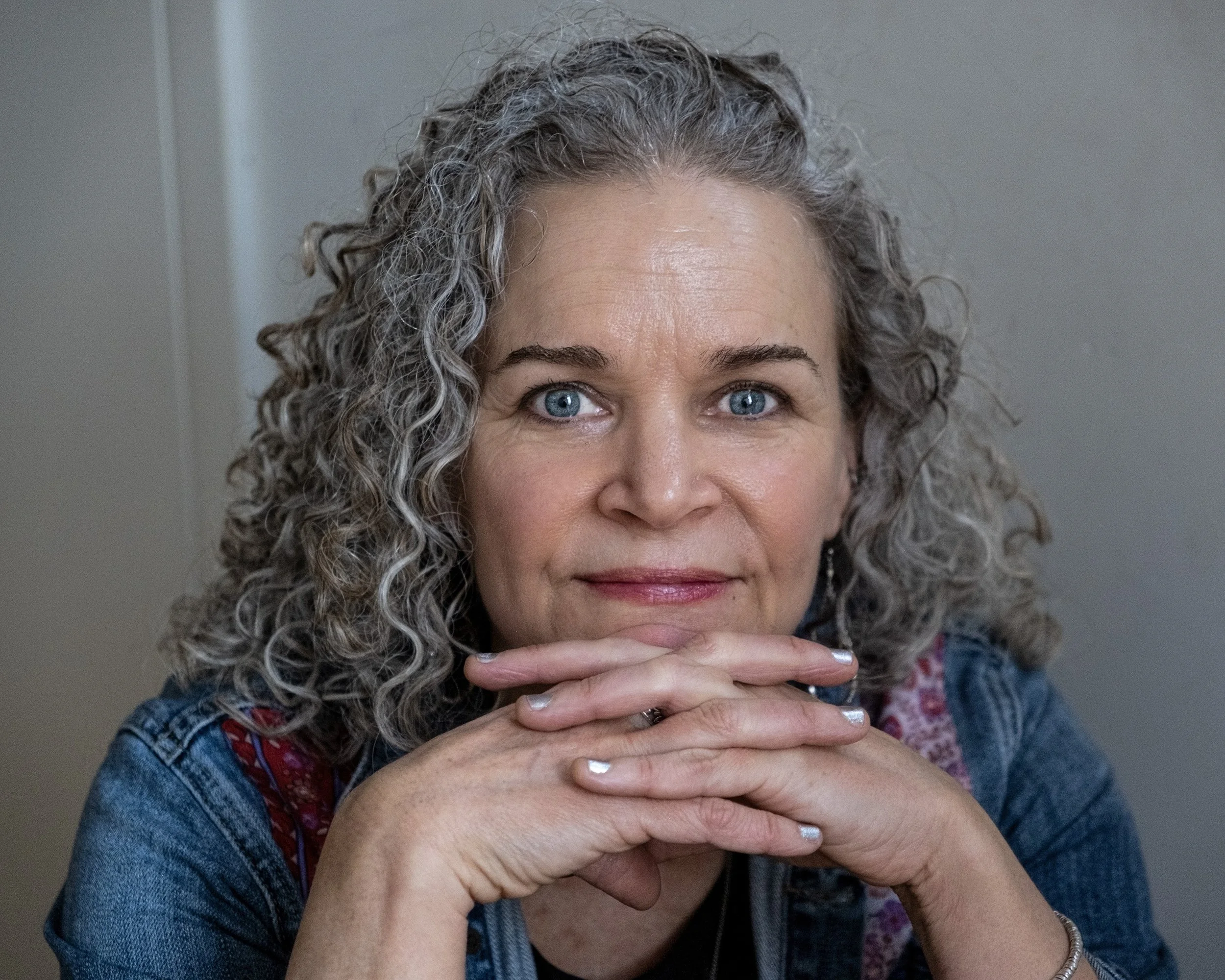 A woman with curly gray hair and blue eyes, resting her chin on her hands, smiling gently at the camera.