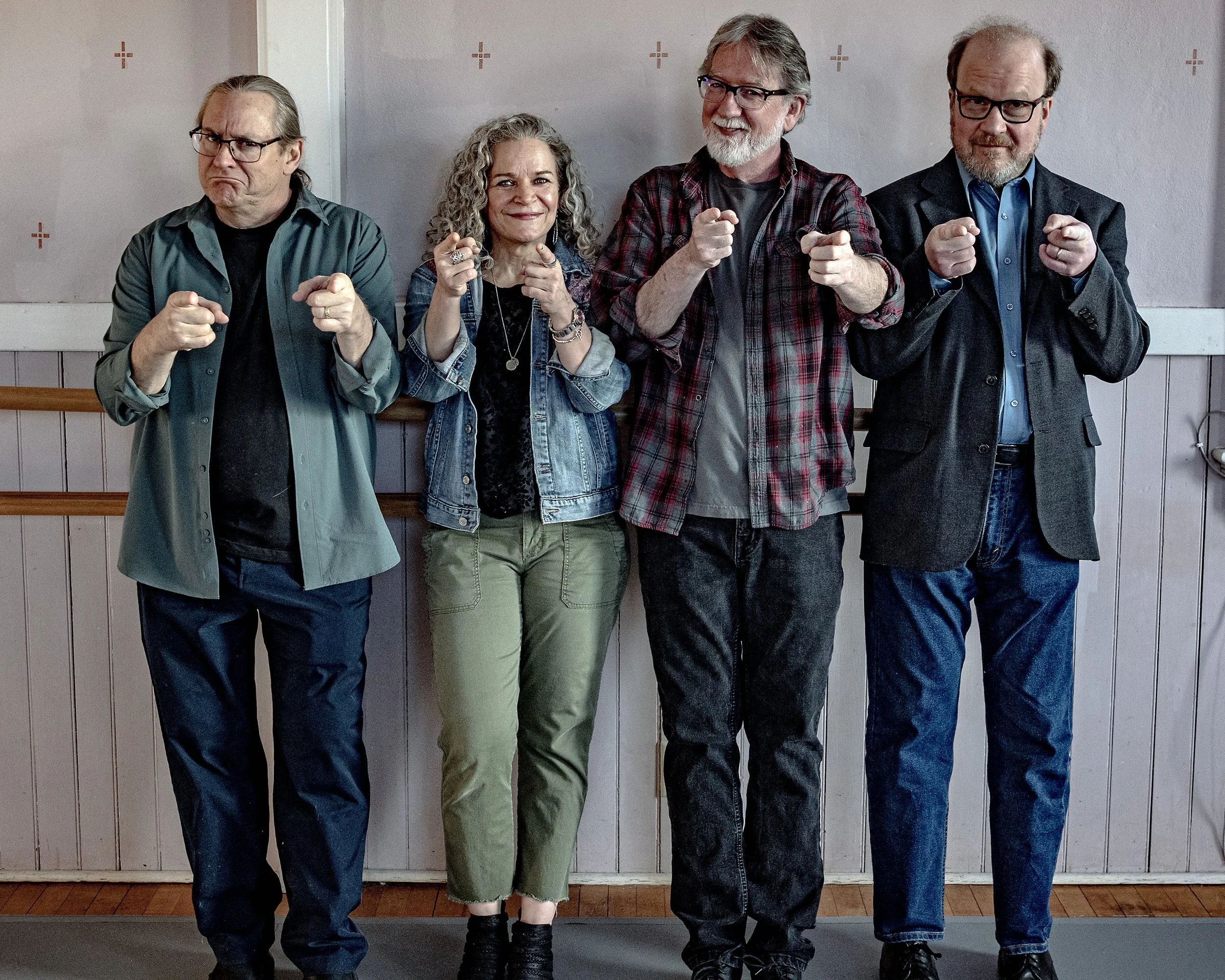 Four adults standing side by side against a wall, pointing forward. They are dressed casually and semi-formally.