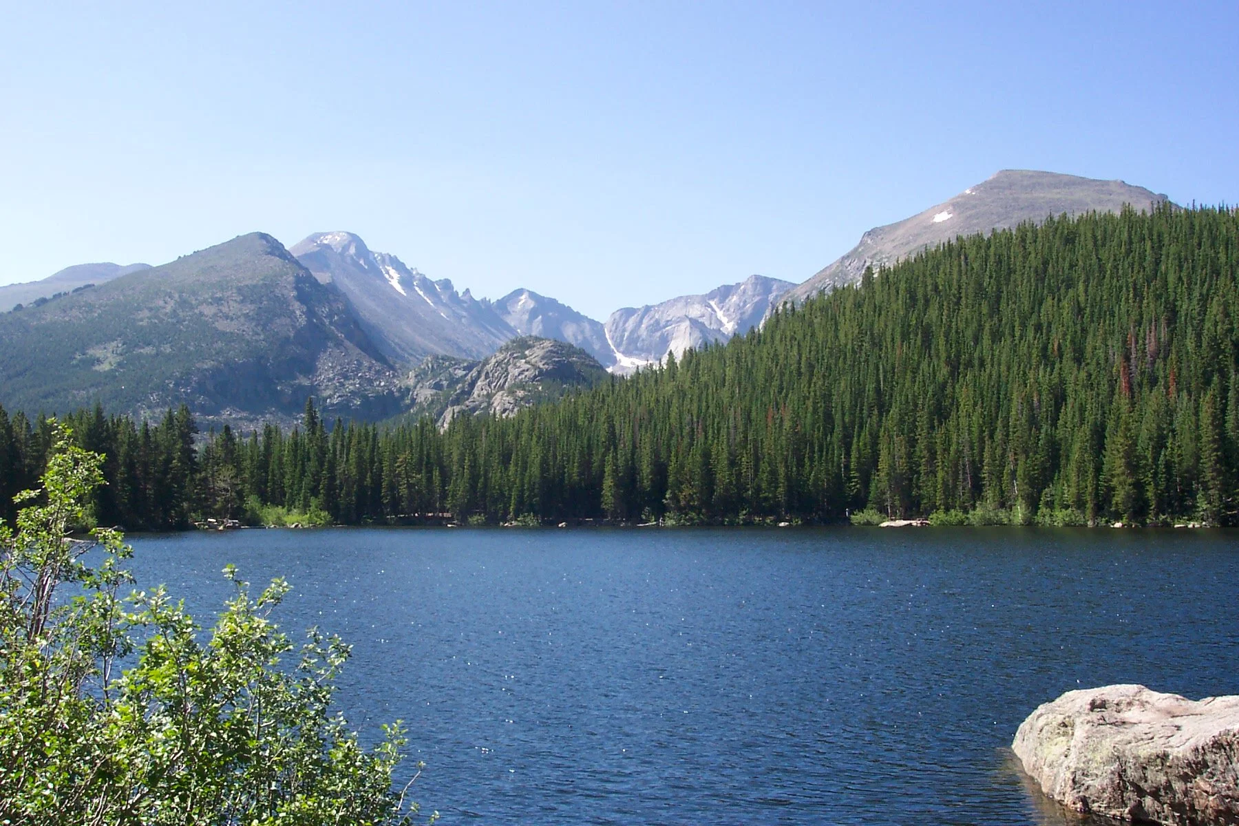 A mountain lake surrounded by dense evergreen forest, with mountains in the background and clear blue sky.